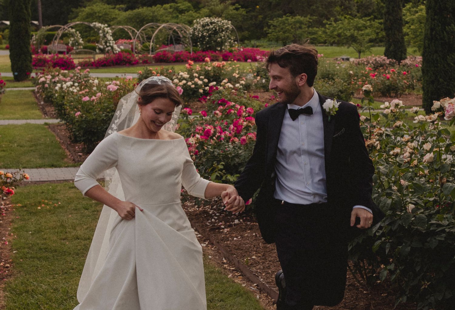 A couple in wedding attire walks together through a garden filled with blooming pink roses and flowering bushes.