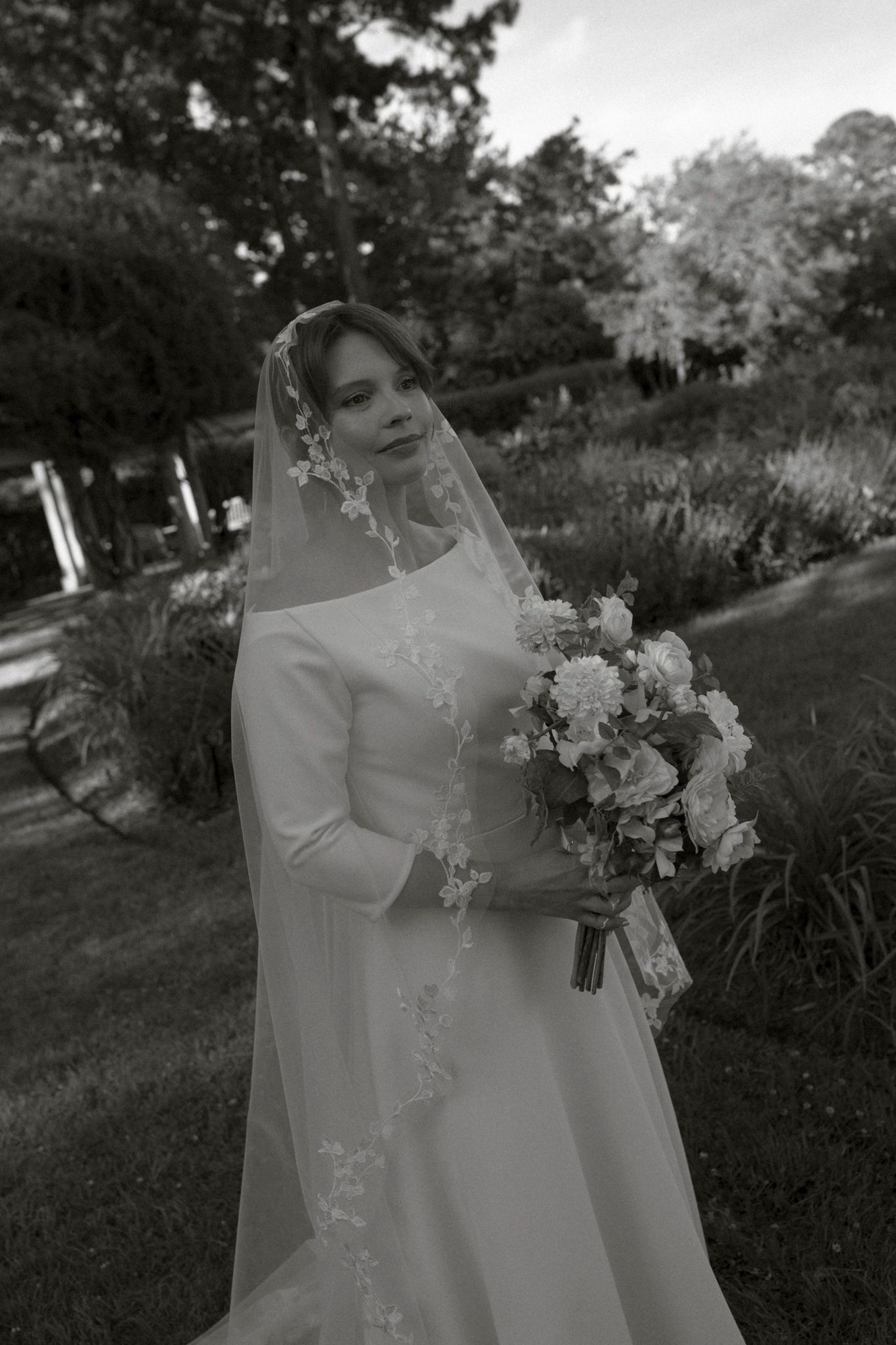 A bride in a flowing white gown and veil holds a delicate floral bouquet in an outdoor garden setting.