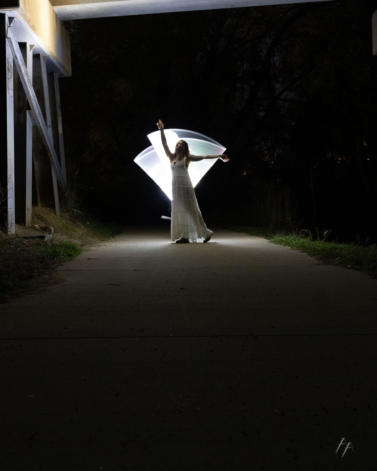 A figure in a white flowing dress creates a striking silhouette against a dark path illuminated by overhead lights.