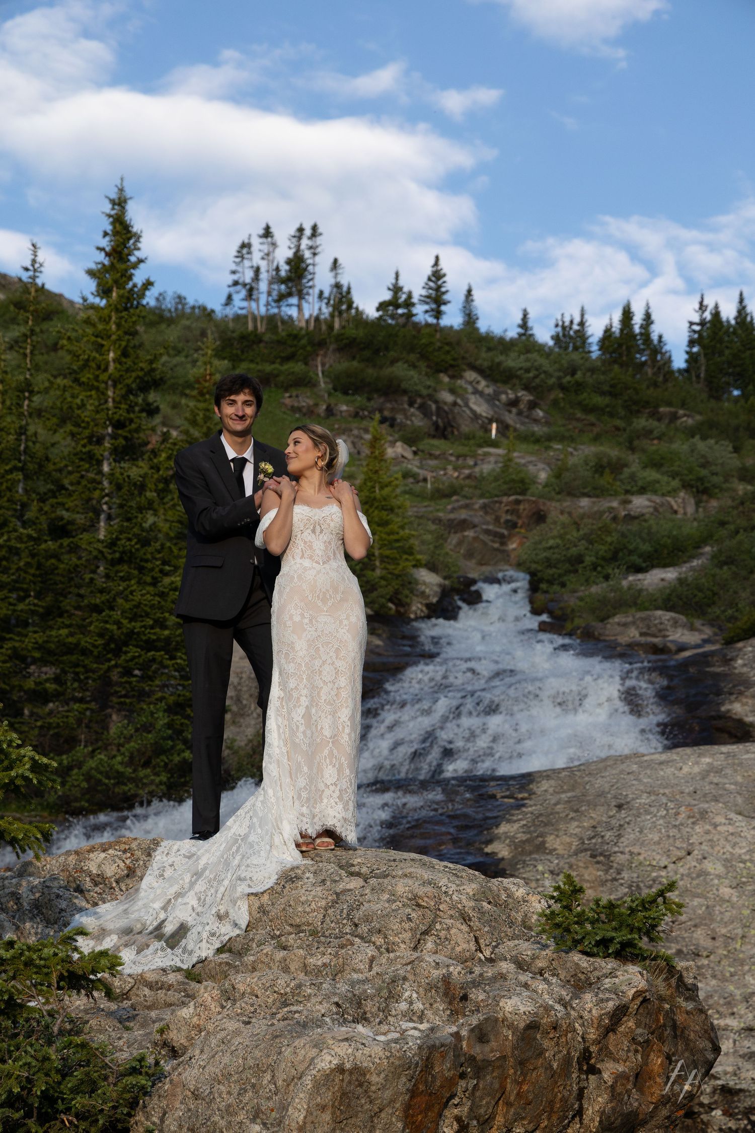 Bride and groom standing on rocky outcrop before a waterfall surrounded by evergreen trees under a blue sky.