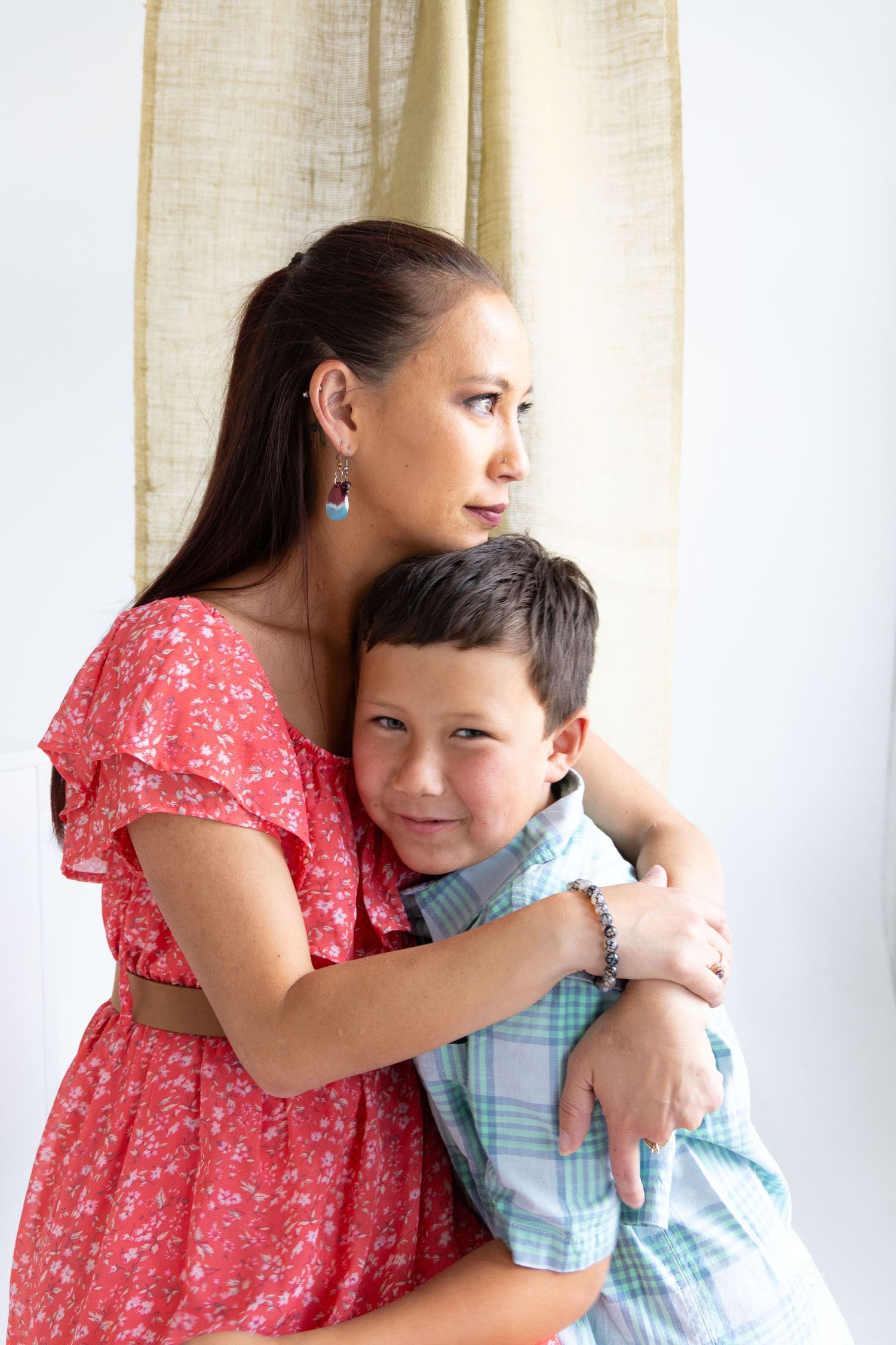A tender embrace between parent and child standing by a window, with the parent wearing a red floral dress.