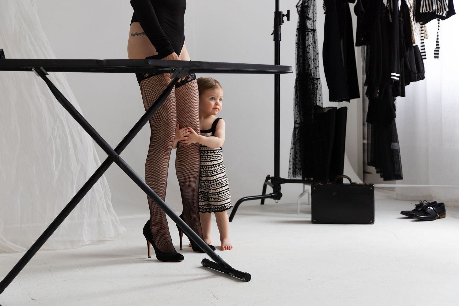 A young child stands beside an ironing board near a woman in black stockings and heels in a bright studio setting.