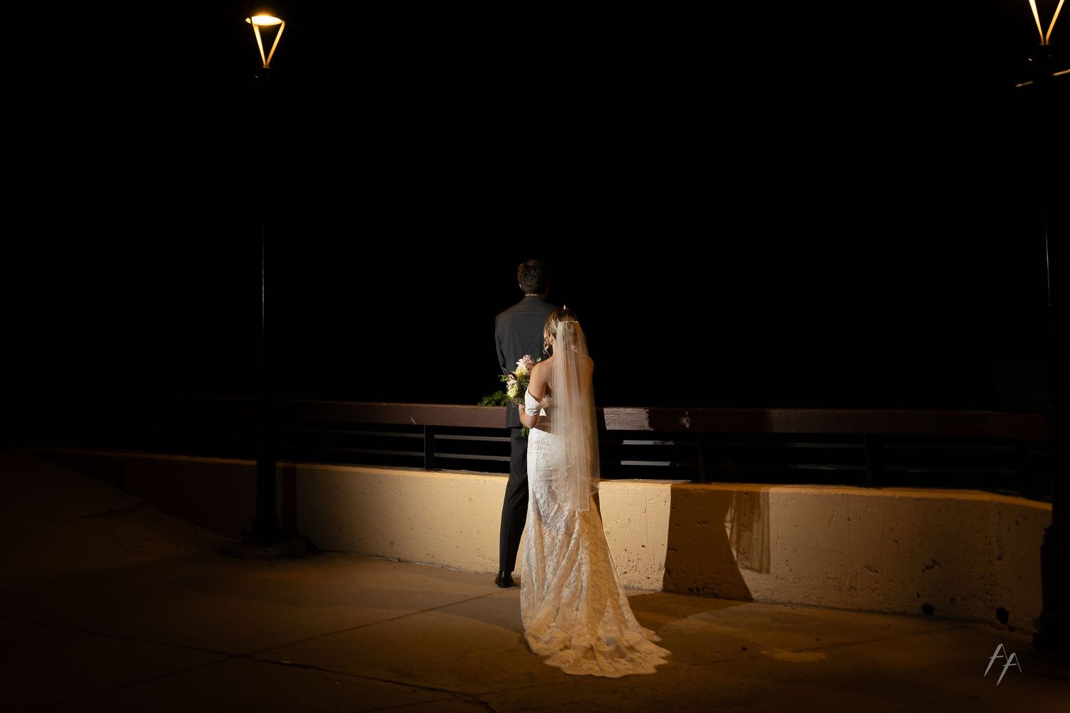 Bride and groom stand together at night under a lamppost, bride wearing a lace gown with veil holding a bouquet.