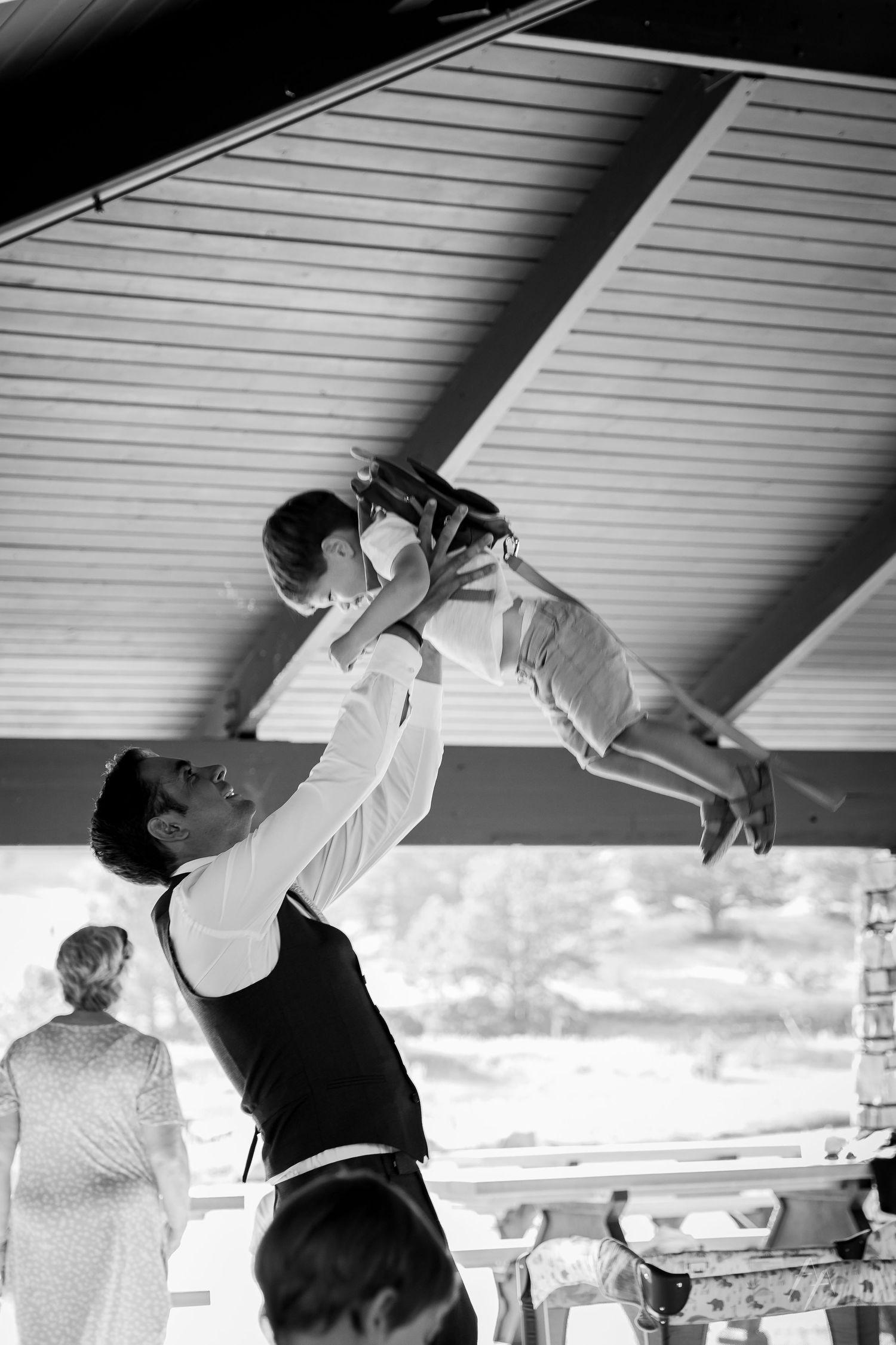 Black and white photo of a groom lifting a young child overhead at a wedding reception under a covered pavilion.