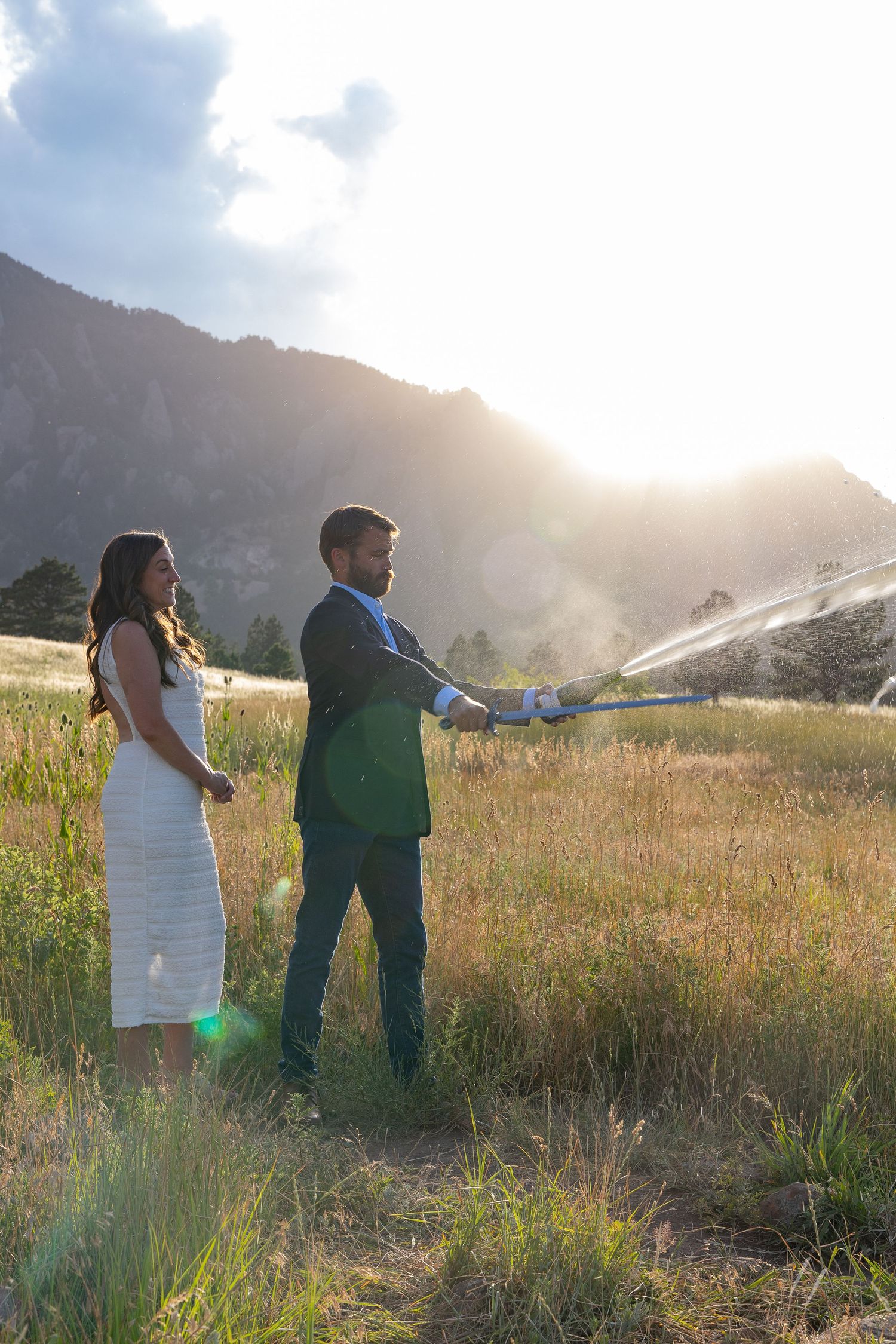 Couple in formal attire using a garden hose in a sunlit mountain meadow at golden hour.
