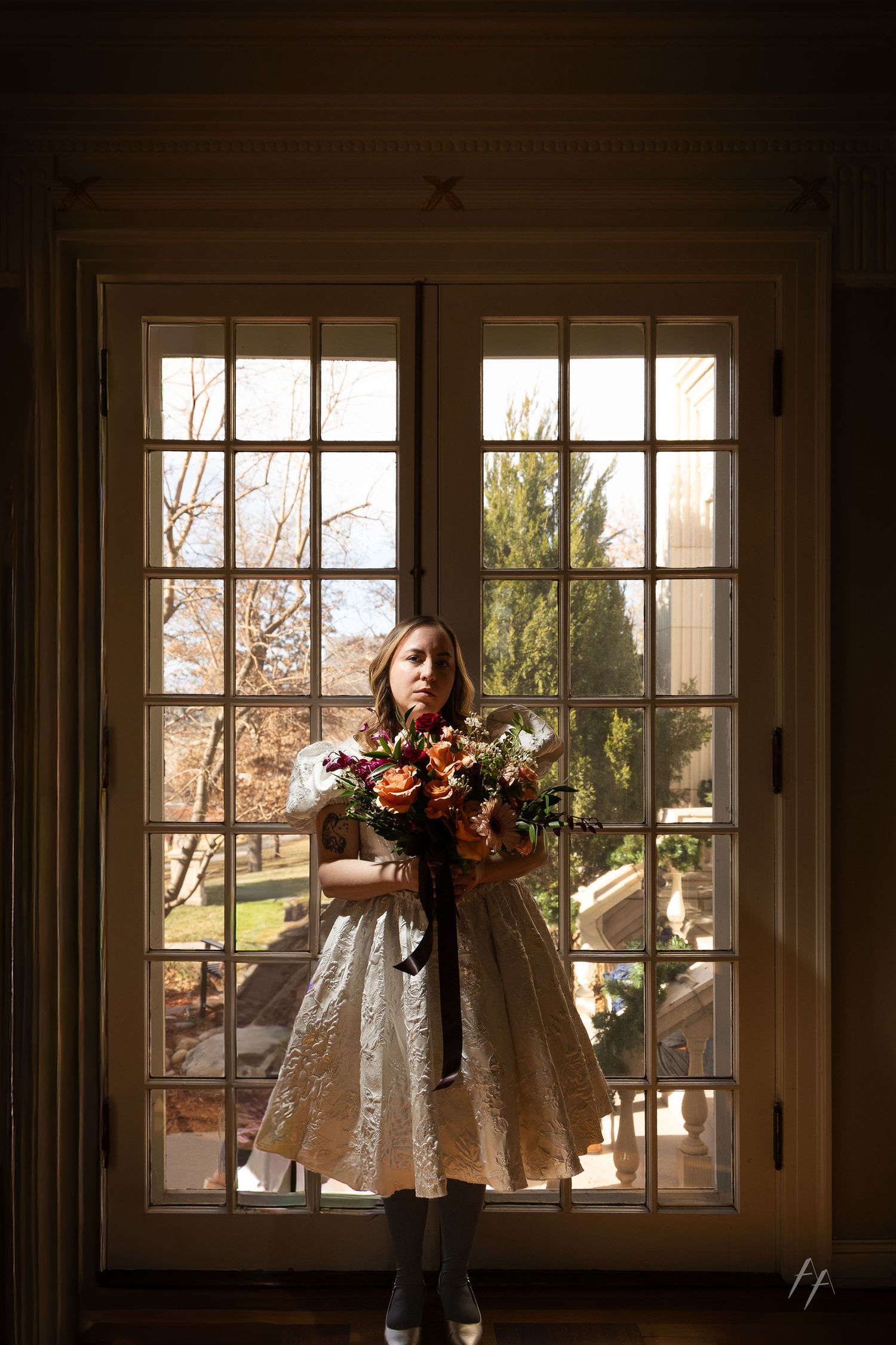 A silhouetted figure in a white dress holds a vibrant autumn bouquet while standing in front of a sunlit French window.