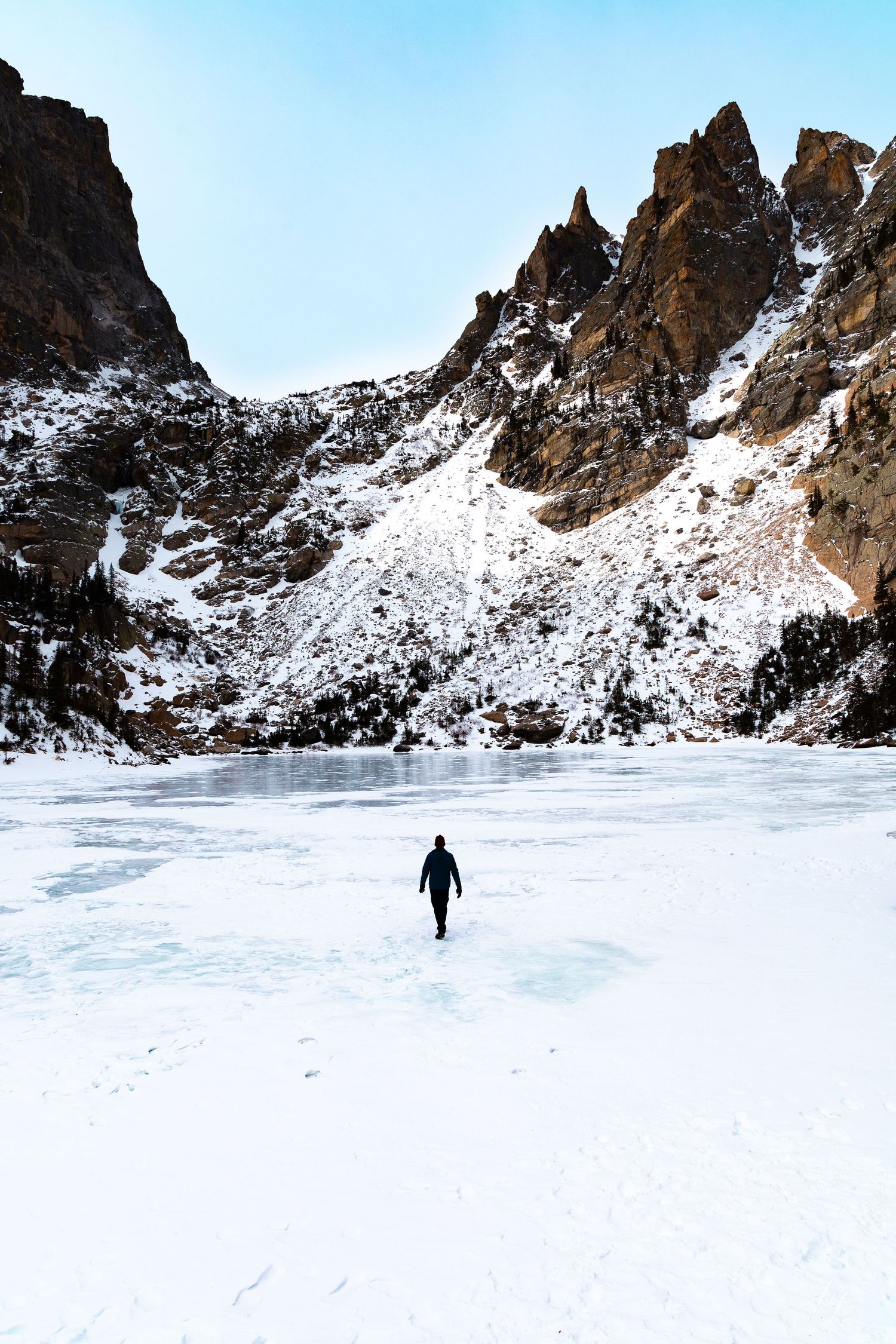 A lone figure walks across a frozen lake beneath towering snow-dusted mountain cliffs. Rocky Mountain National Park