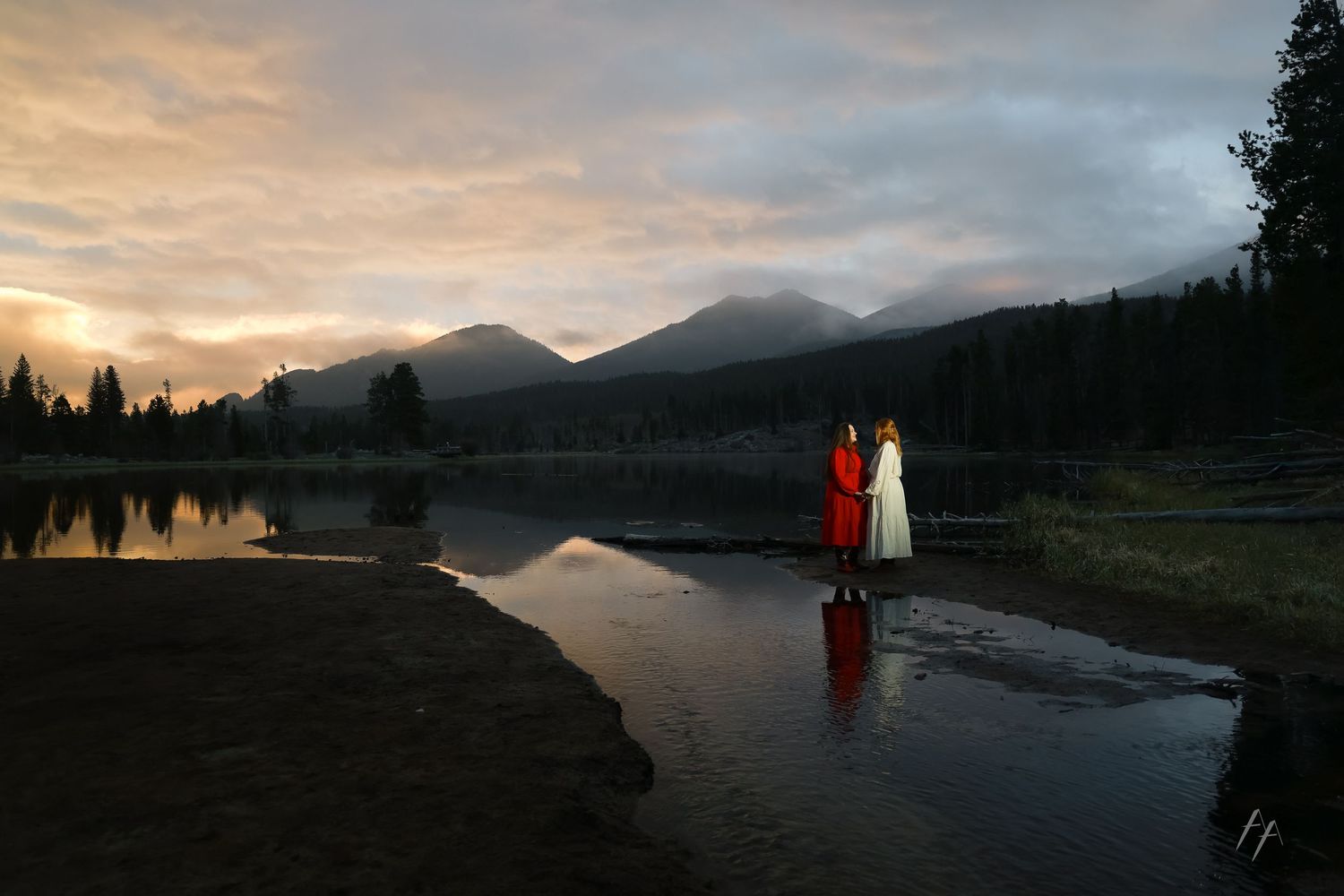 Two people in colorful robes stand by a reflective mountain lake at sunset with dramatic clouds and forested peaks.
