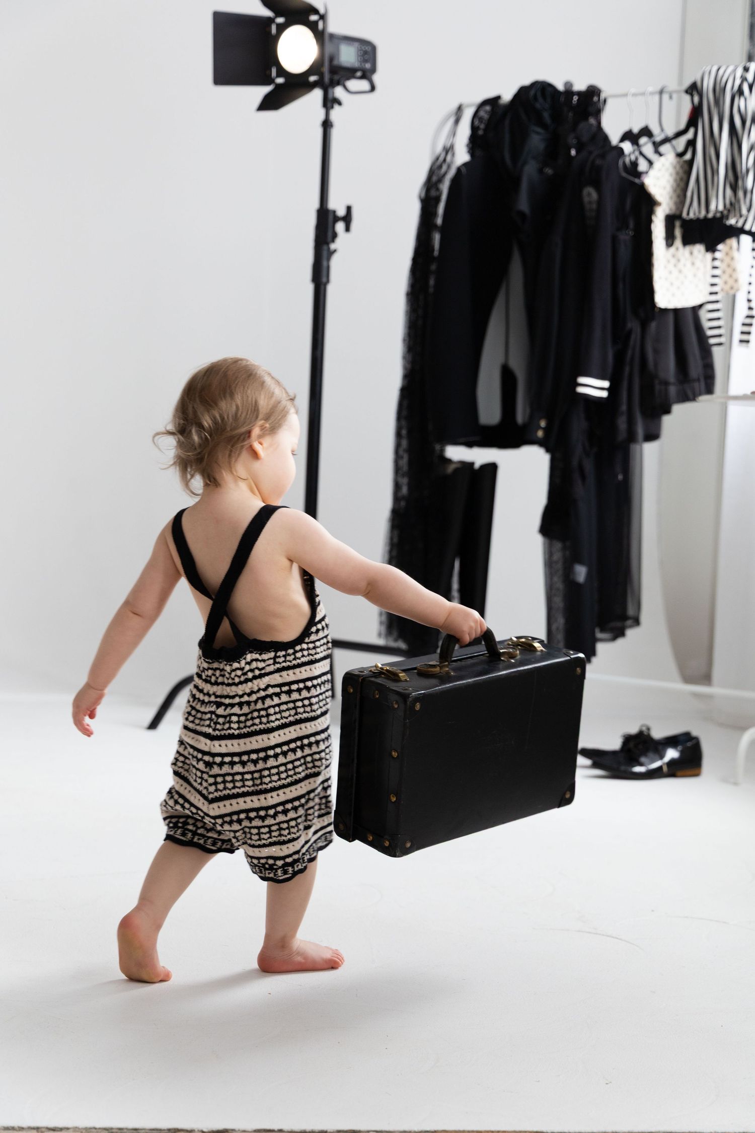 Toddler in patterned romper carrying a black suitcase in a photography studio with clothing rack.