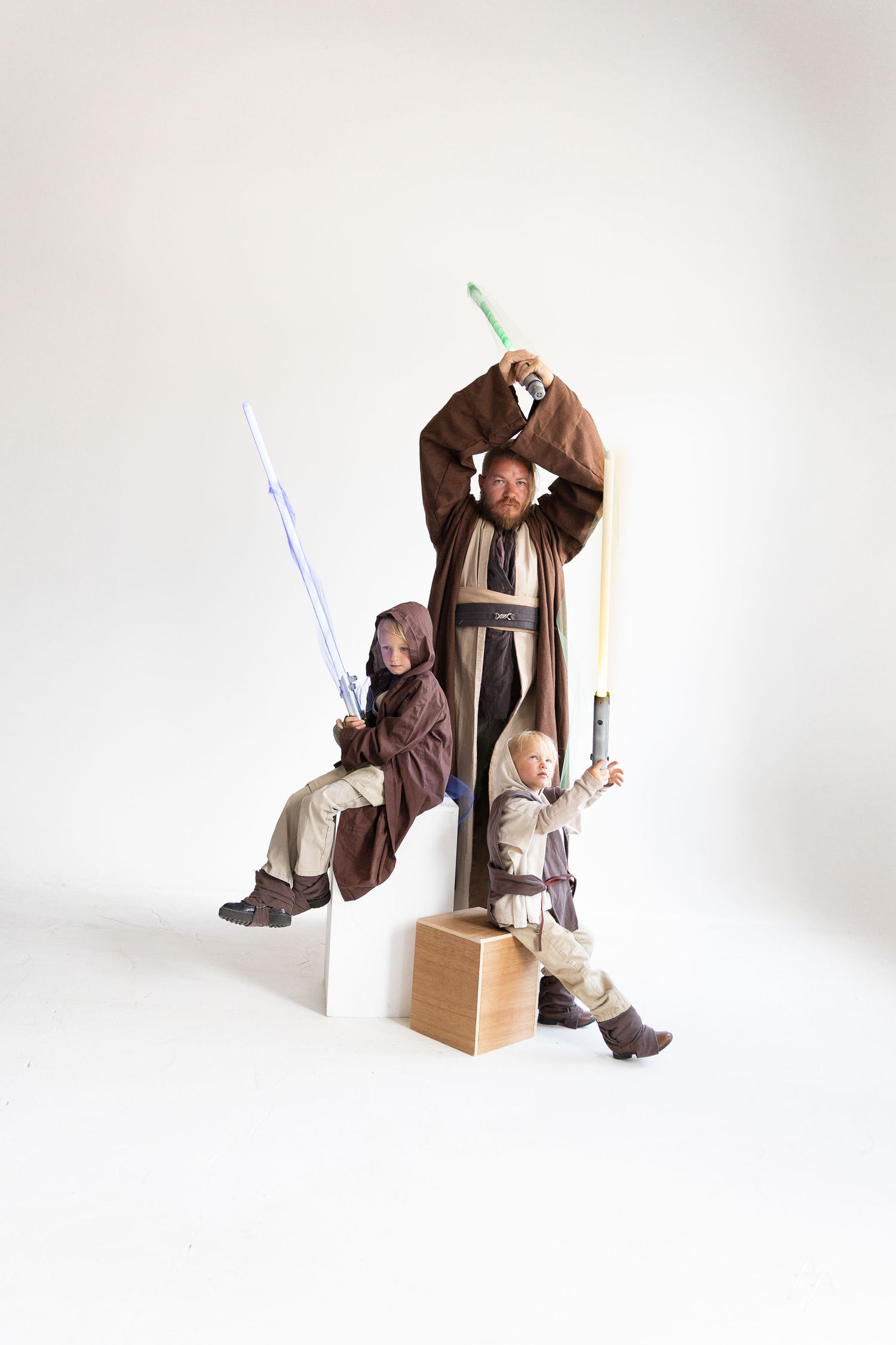 Three people dressed in brown Jedi robes pose with lightsabers against a white background in a Star Wars themed photo.