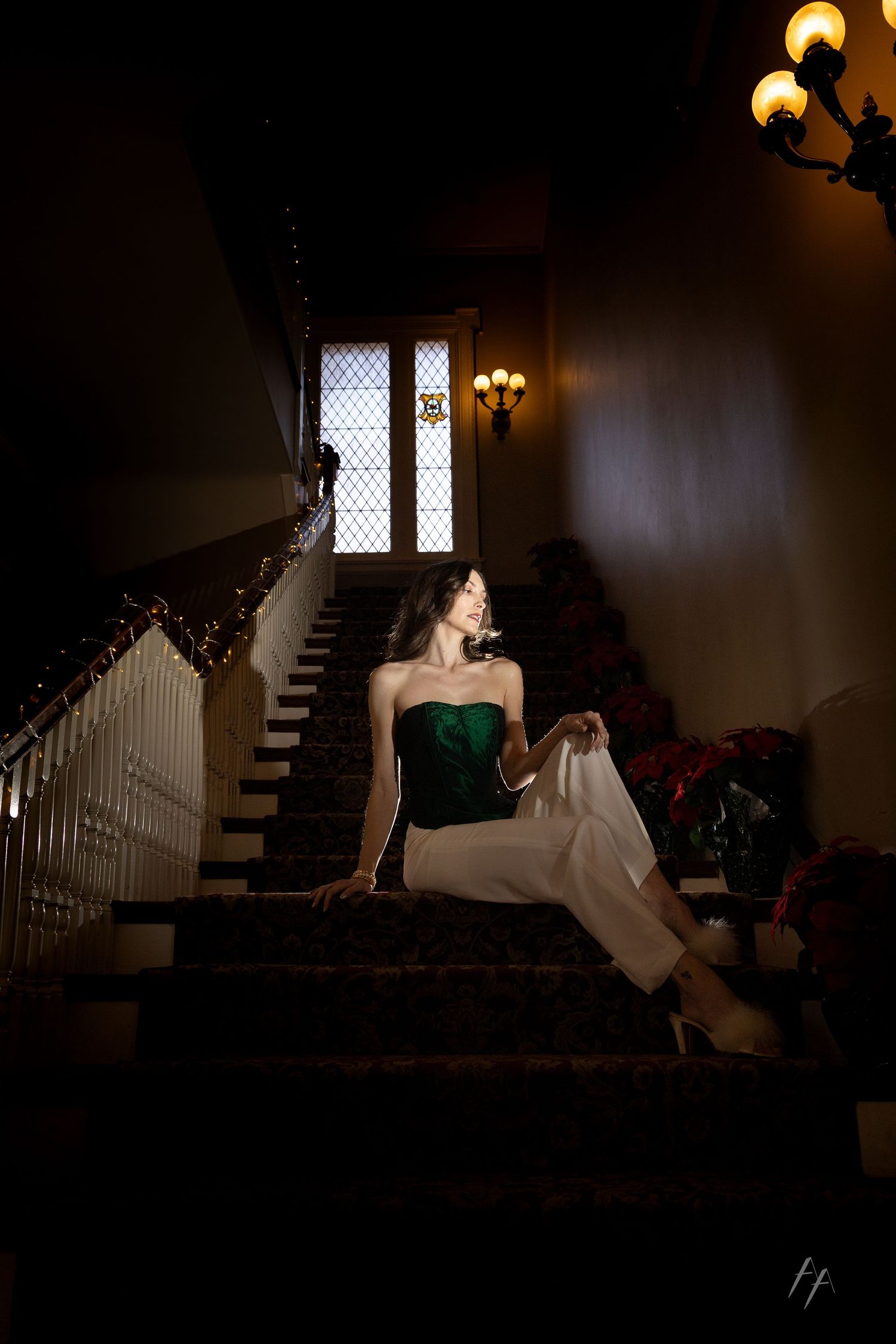 Elegant figure in emerald green dress poses dramatically on dark staircase beneath gothic window and vintage lighting.