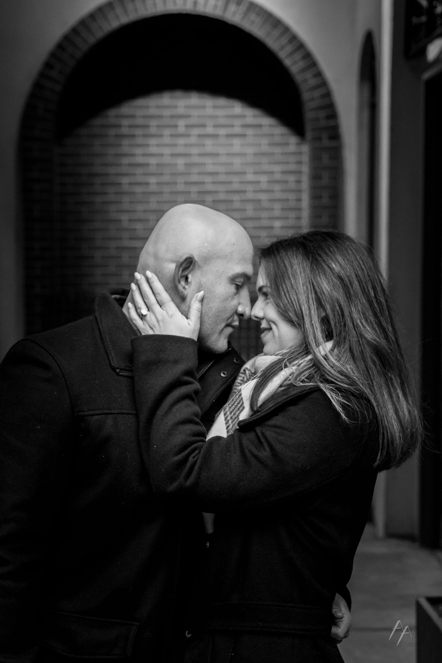 Black and white photo of a couple sharing a romantic moment, foreheads touching, with a brick arch backdrop.
