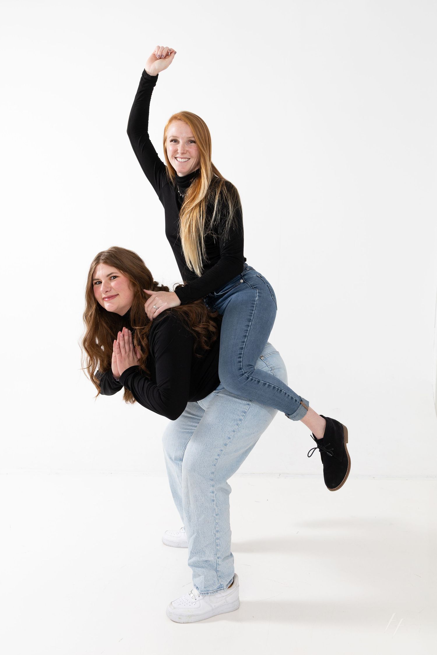 Two people in black tops and jeans pose playfully against white background with one person giving a piggyback ride.