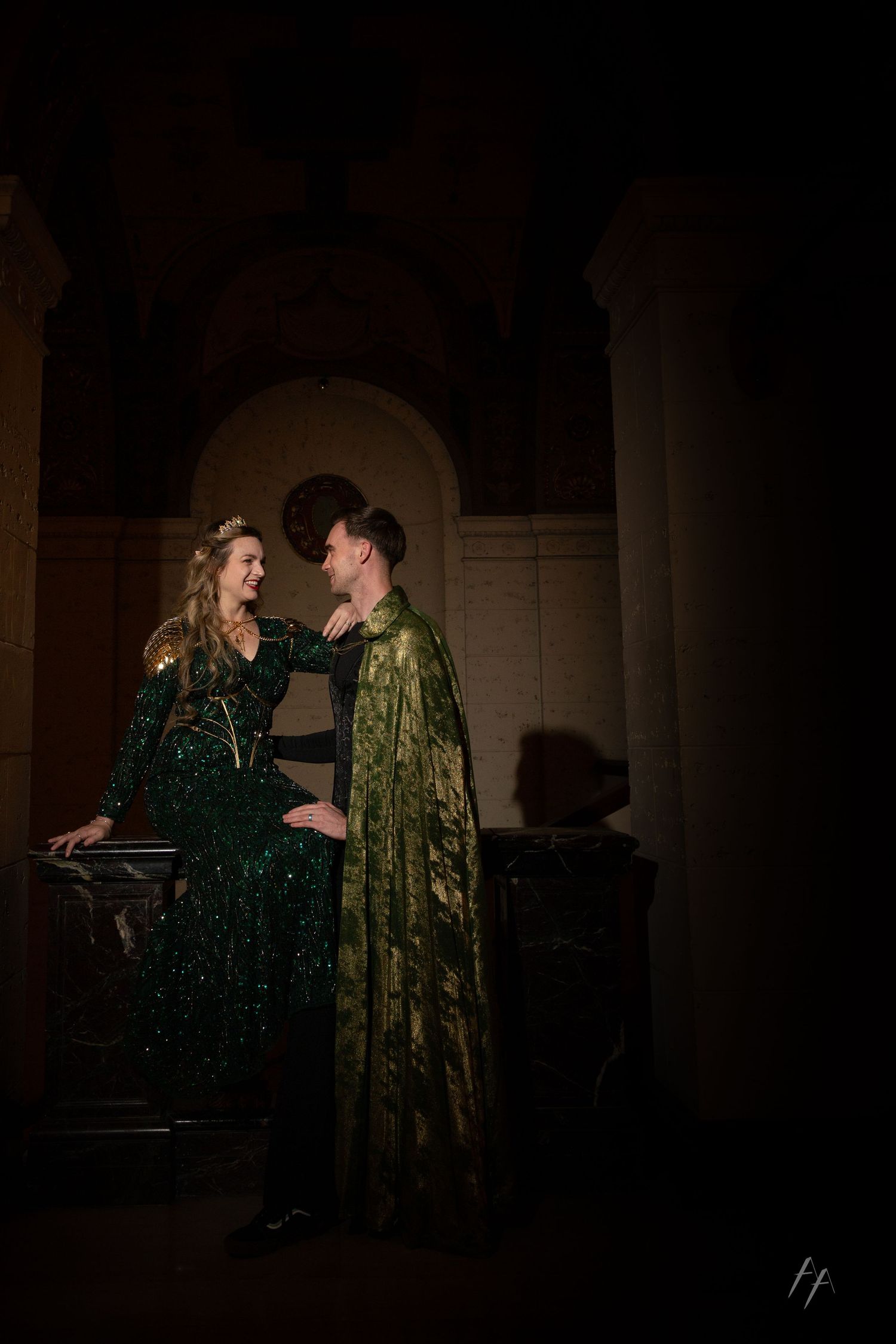 Two performers in green sequined and velvet costumes interact dramatically on a dimly lit stage during a theatrical scene.