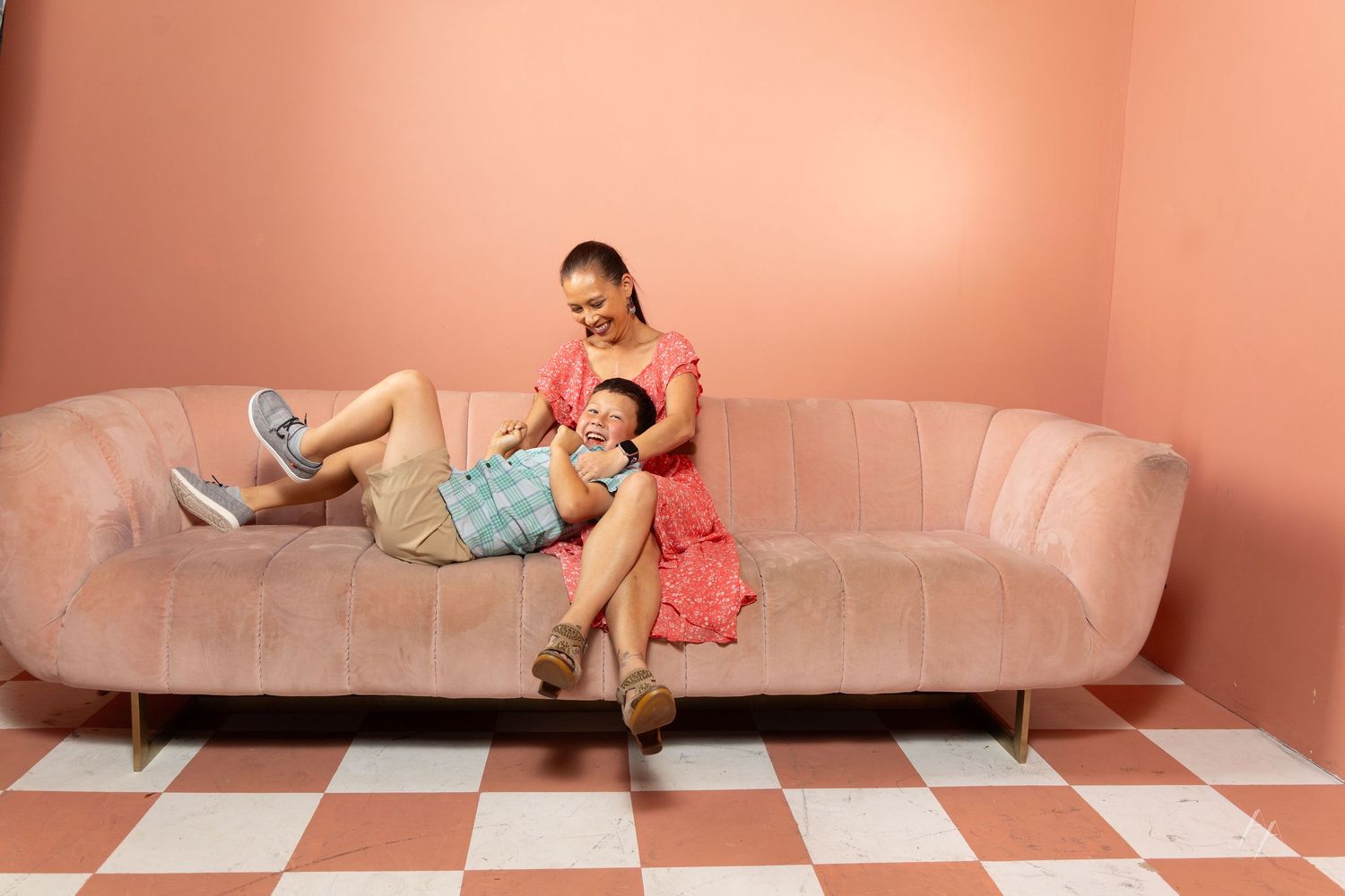 Woman in pink floral dress sits on blush velvet sofa with child in checkered room with pink walls.