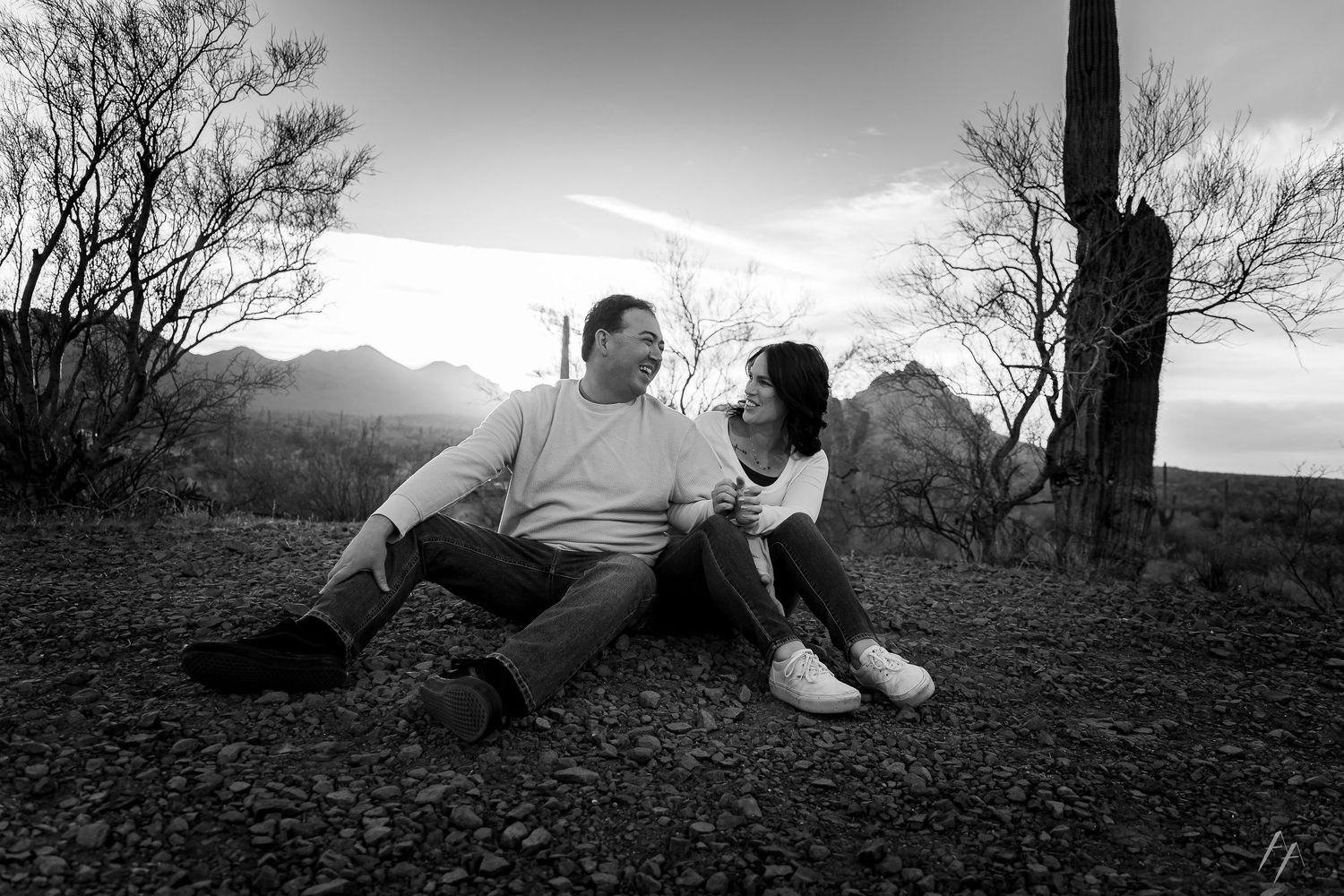 Black and white photo of a couple sitting together on rocky ground with mountains and bare trees in the background.