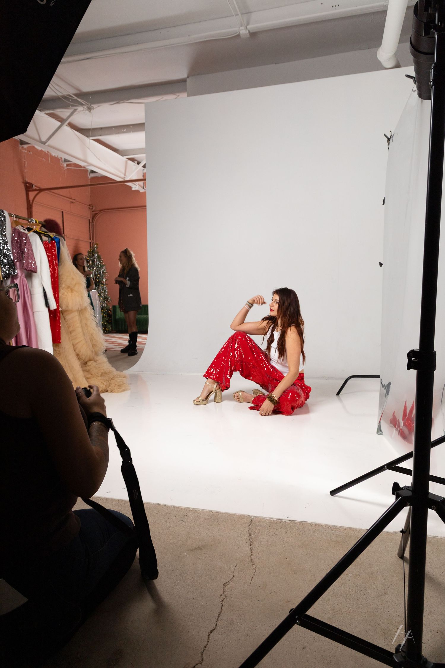 Woman in red outfit poses on white backdrop in professional photo studio with lighting equipment and clothing rack.