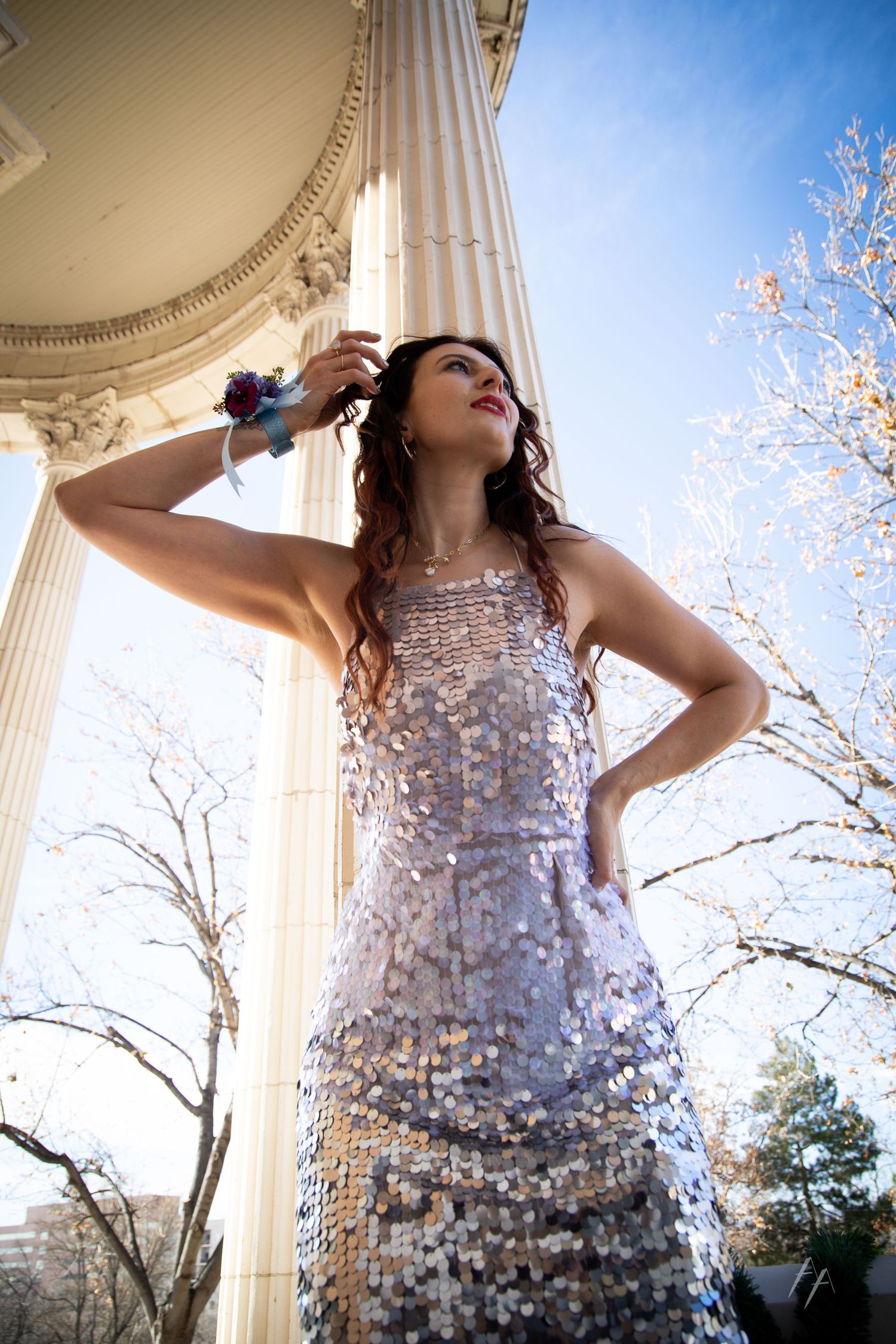 A figure in a shimmering dress poses dramatically against white columns and a blue sky with blooming trees.