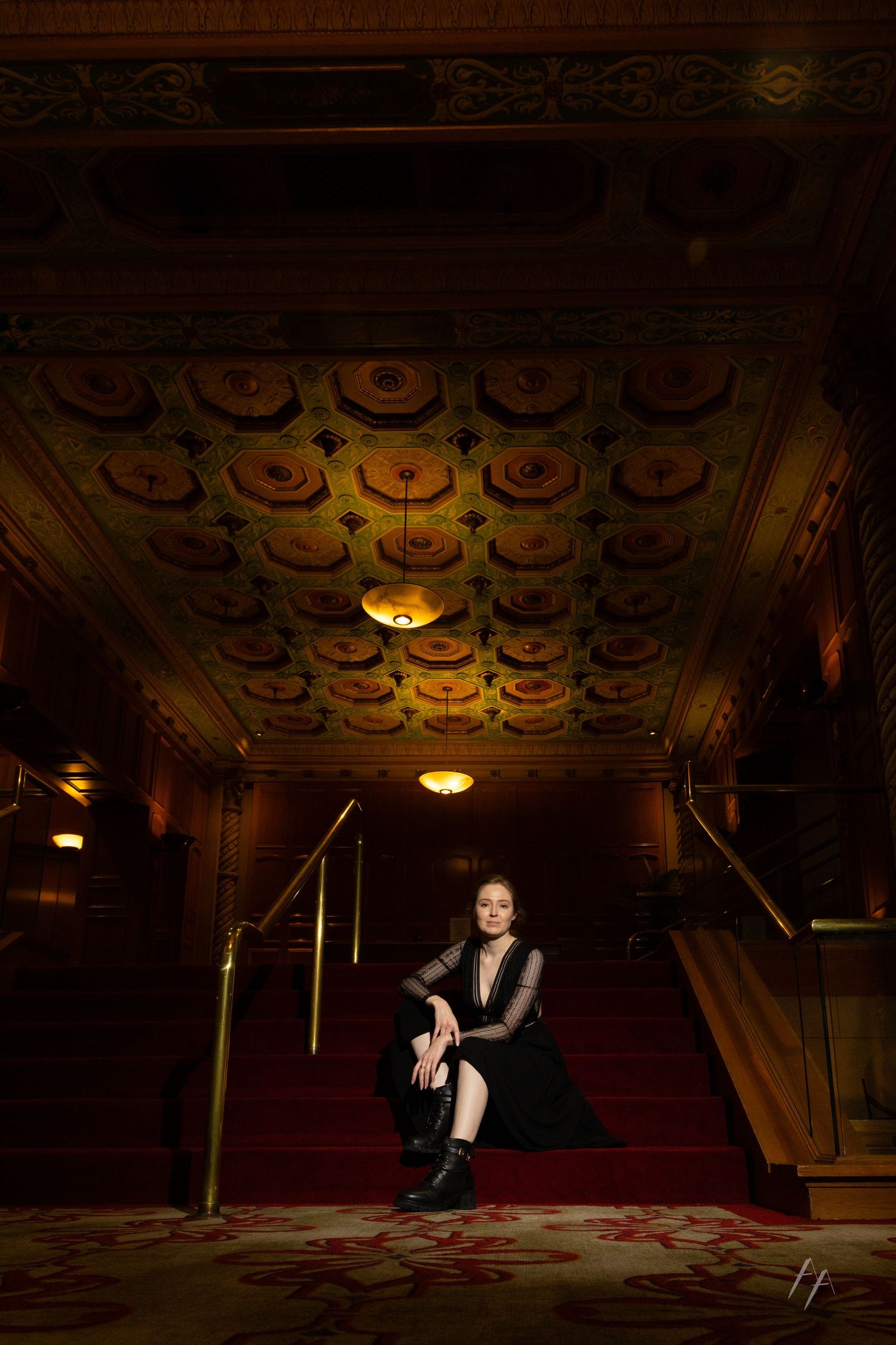 A figure emerges from shadows in a dimly lit ornate theater with decorative ceiling and red carpeting.