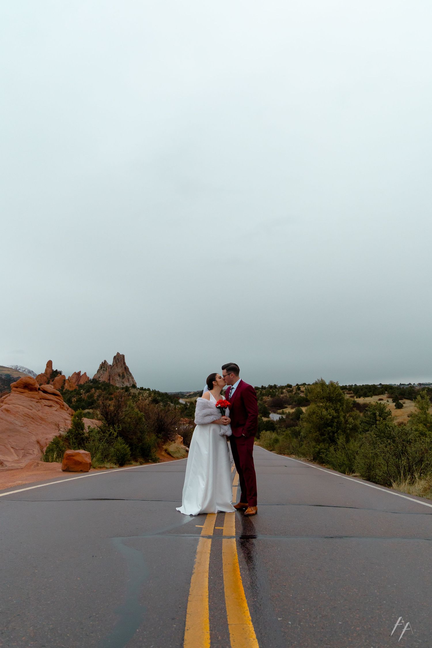A couple shares a romantic moment on a desert road with red rock formations and desert vegetation in the background.