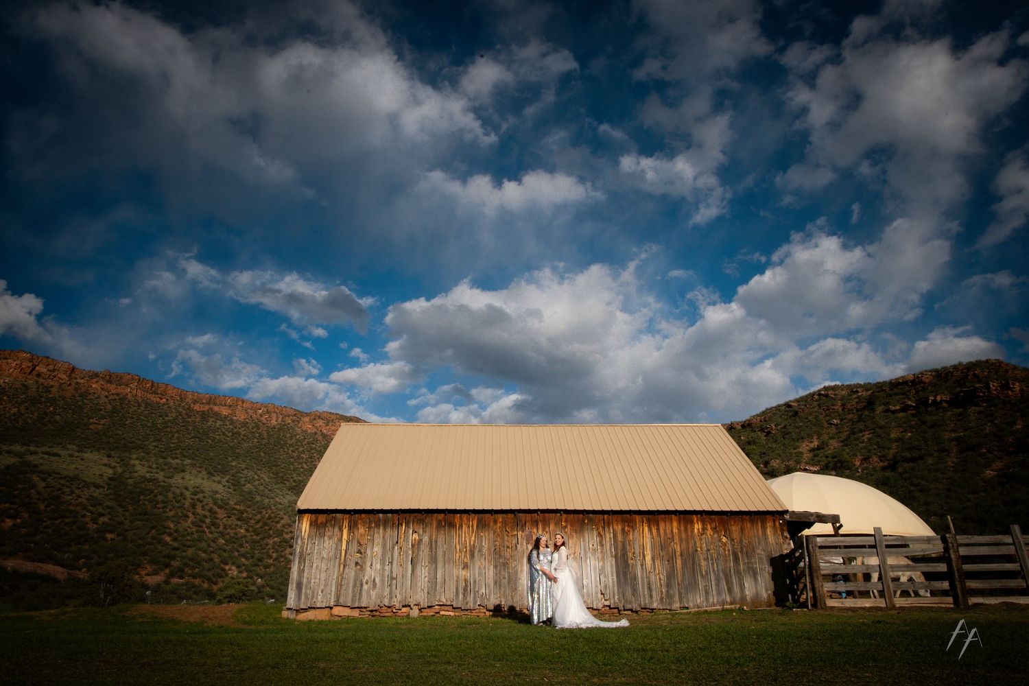 Bride and groom embrace before a rustic barn under dramatic blue skies at a countryside wedding venue.