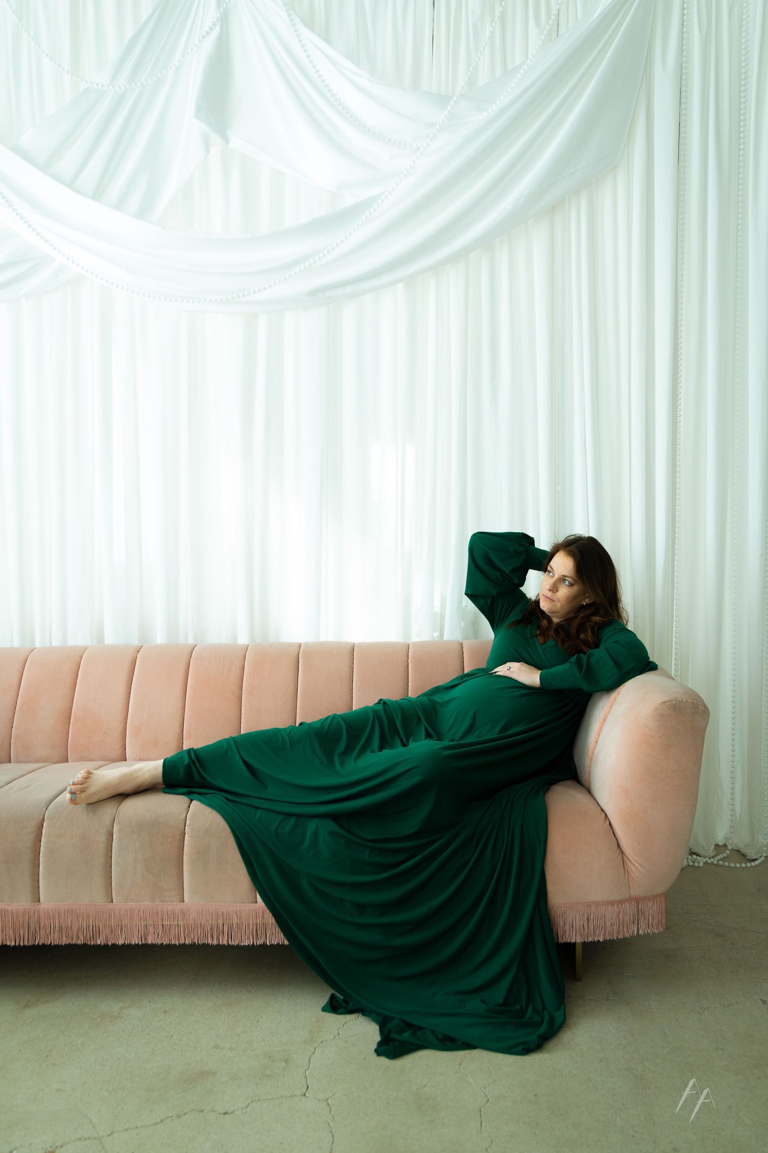 A figure in an emerald green dress reclines dramatically across a pink couch against a white curtained backdrop.