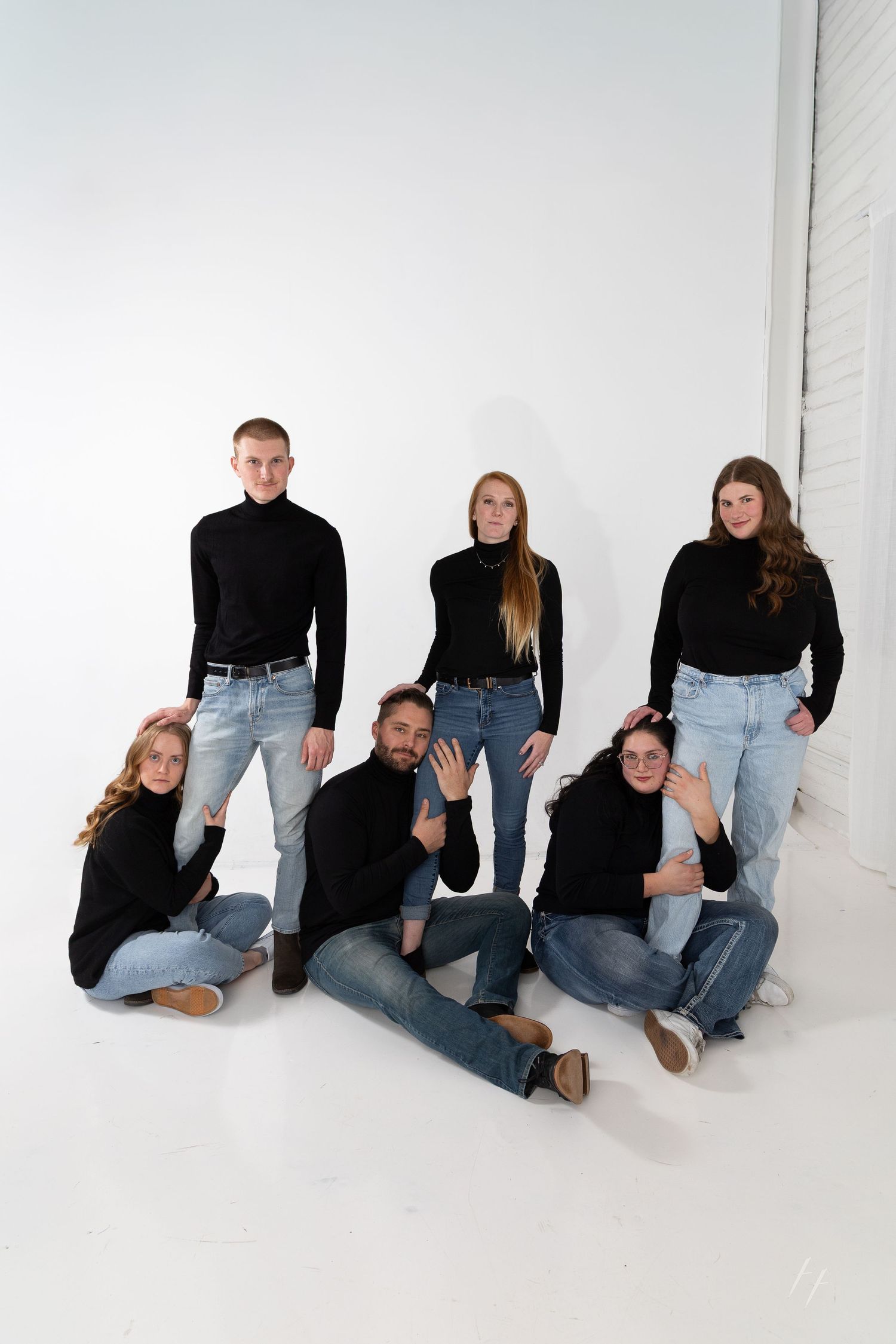 Group of six young adults in matching black turtlenecks and jeans posing together against a white studio background.
