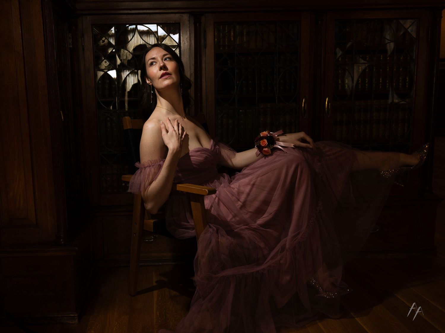 A figure in a flowing mauve dress poses dramatically in moody lighting against a dark wooden interior setting.