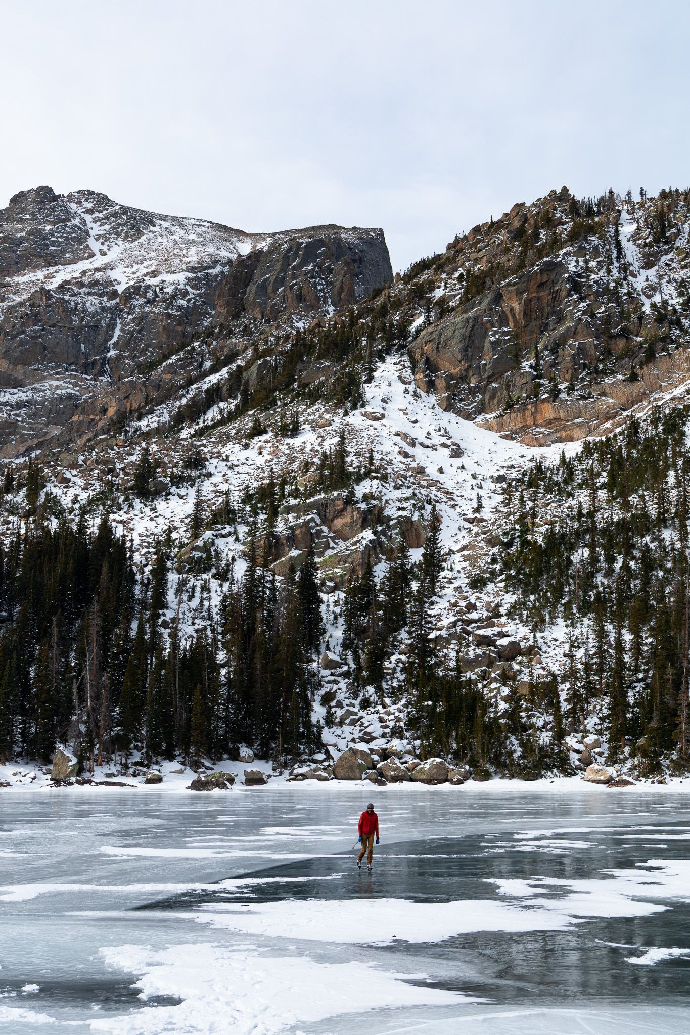 A solitary figure in red stands on a frozen alpine lake surrounded by snow-covered mountain peaks and evergreen forest. Rocky Mountain Natio