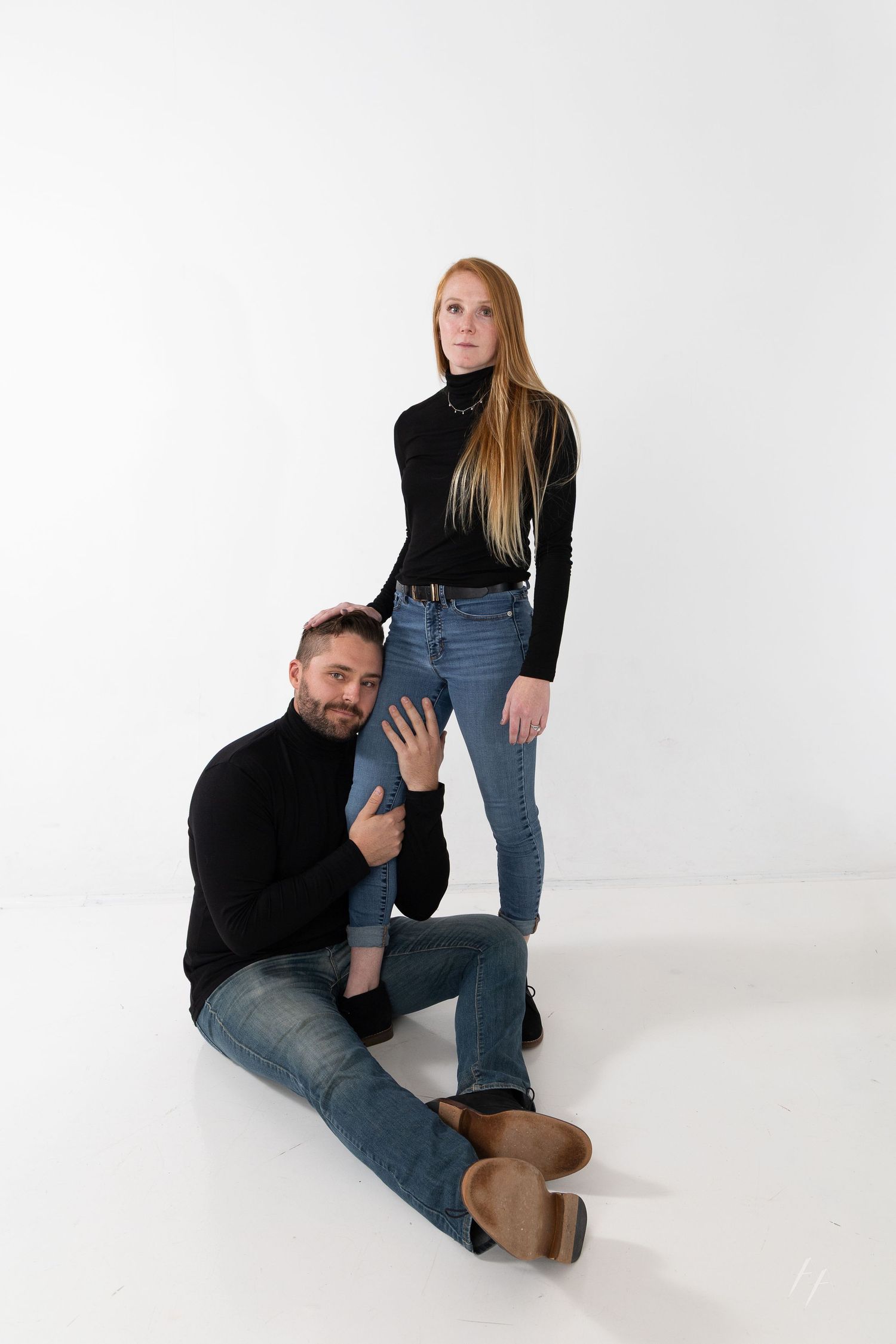 A man and woman in matching black turtlenecks and jeans pose together in a white studio setting.