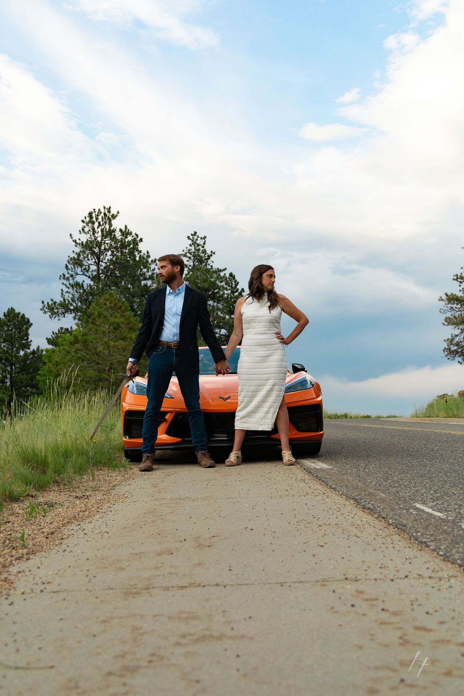 Couple in formal attire posing beside an orange sports car on a rural roadside with trees and cloudy sky.