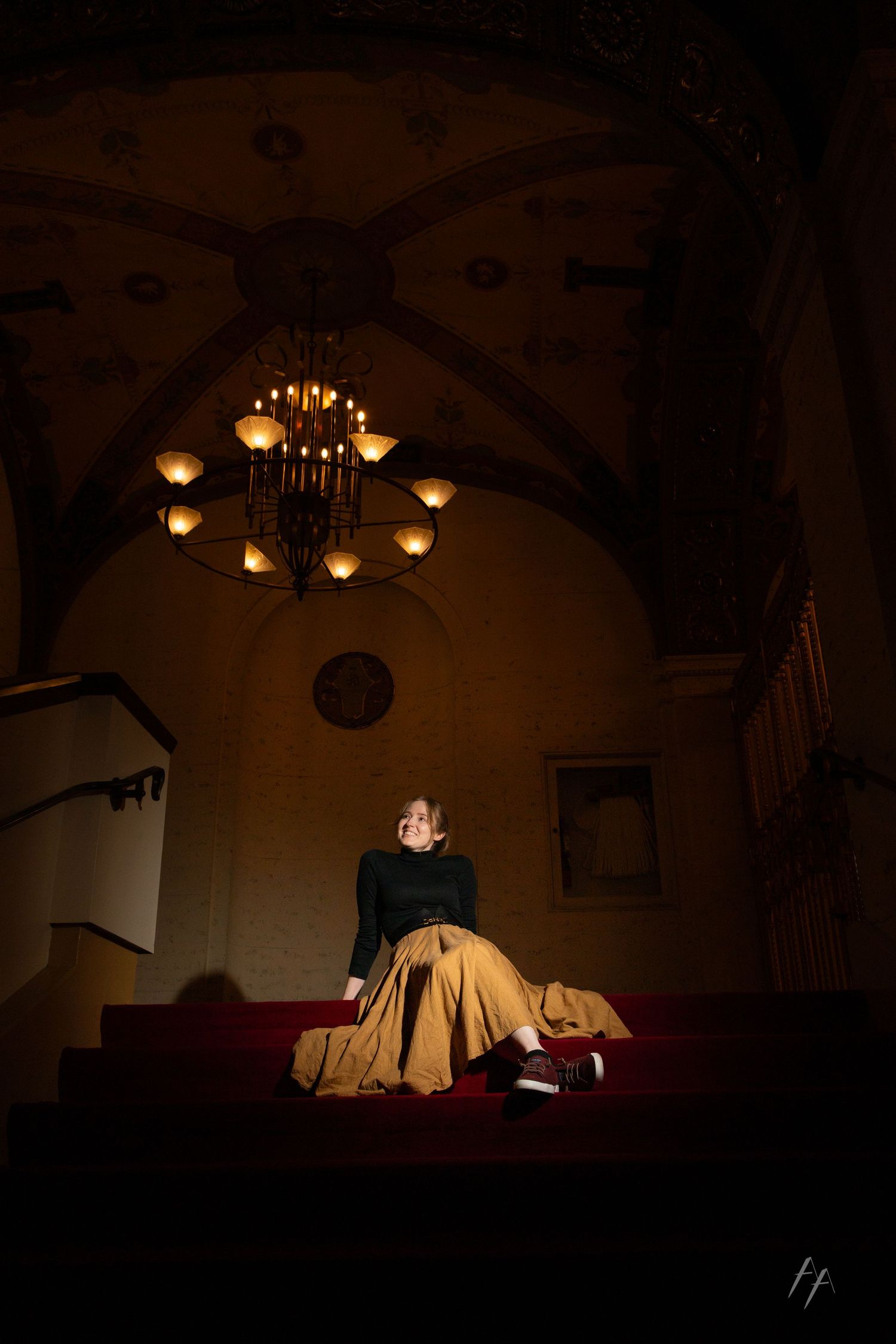 A figure in a black top and beige skirt sits on red steps under a dimly lit chandelier in a dark, dramatic interior.