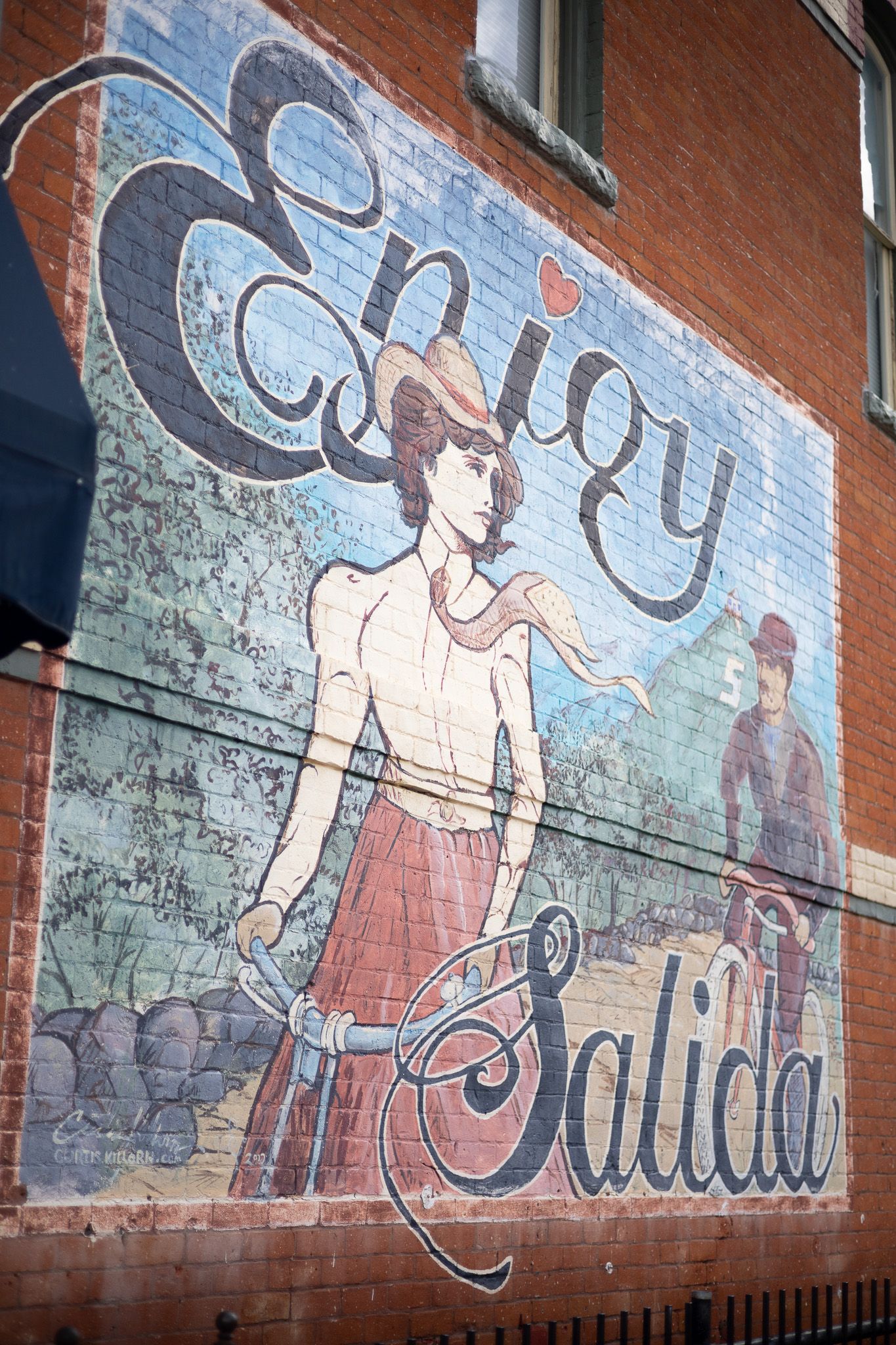 Colorful mural on brick wall reading Enjoy Salida featuring a woman on a bicycle in vintage style artwork.