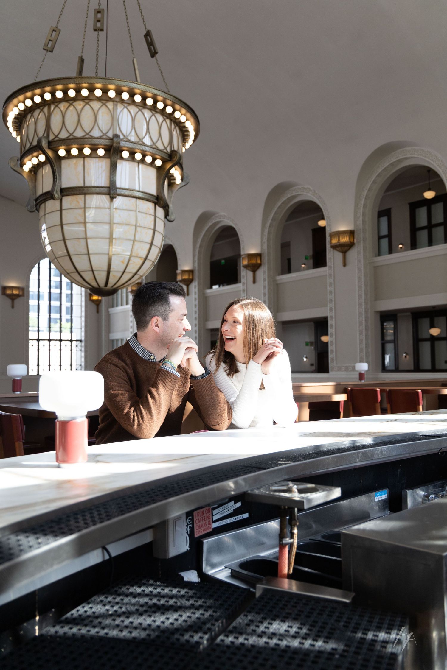 A couple sits at a long table in an elegant room with ornate chandeliers and arched windows.