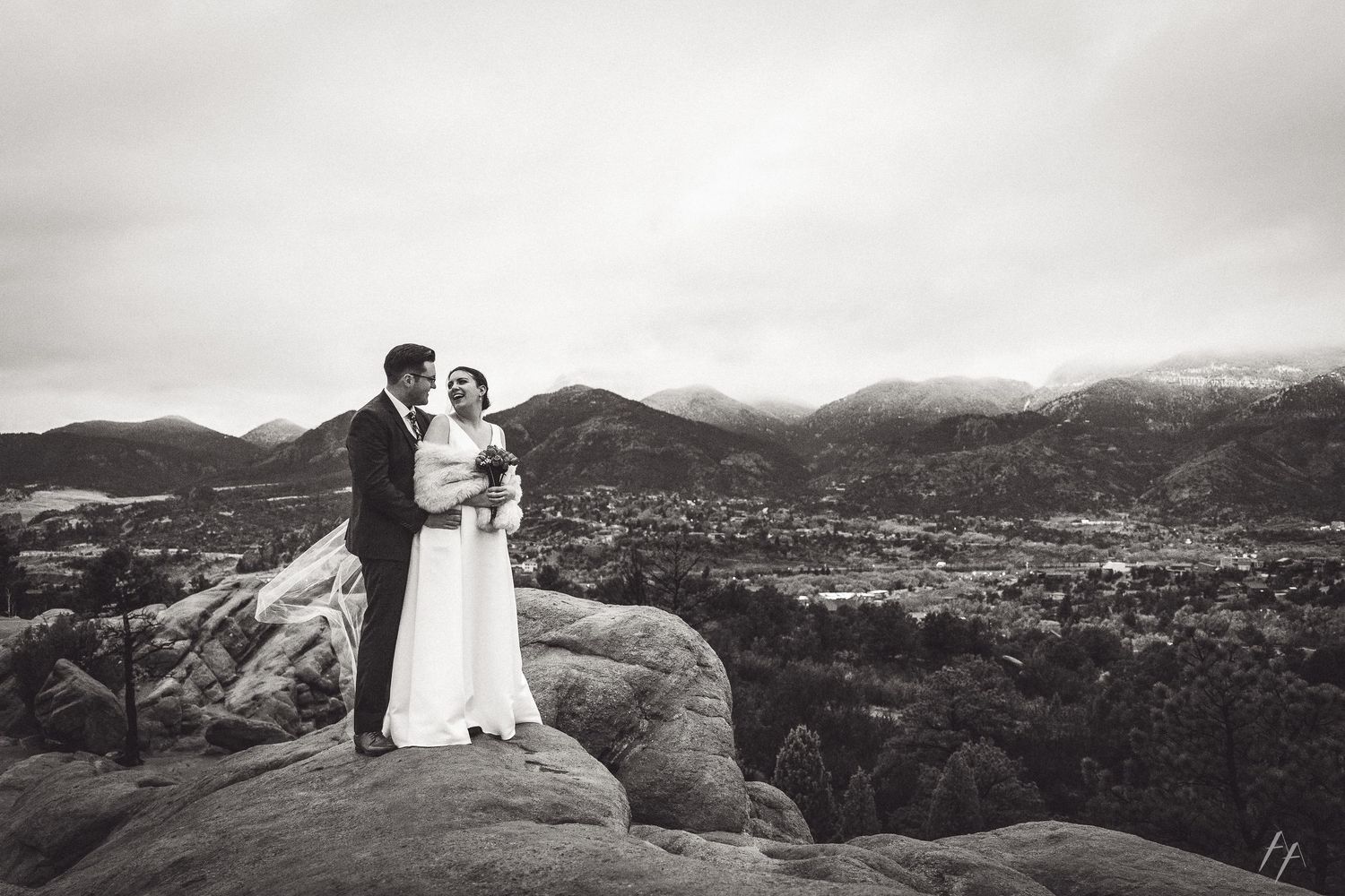 Couple in wedding attire poses on mountain ridge overlooking dramatic desert landscape at sunset.