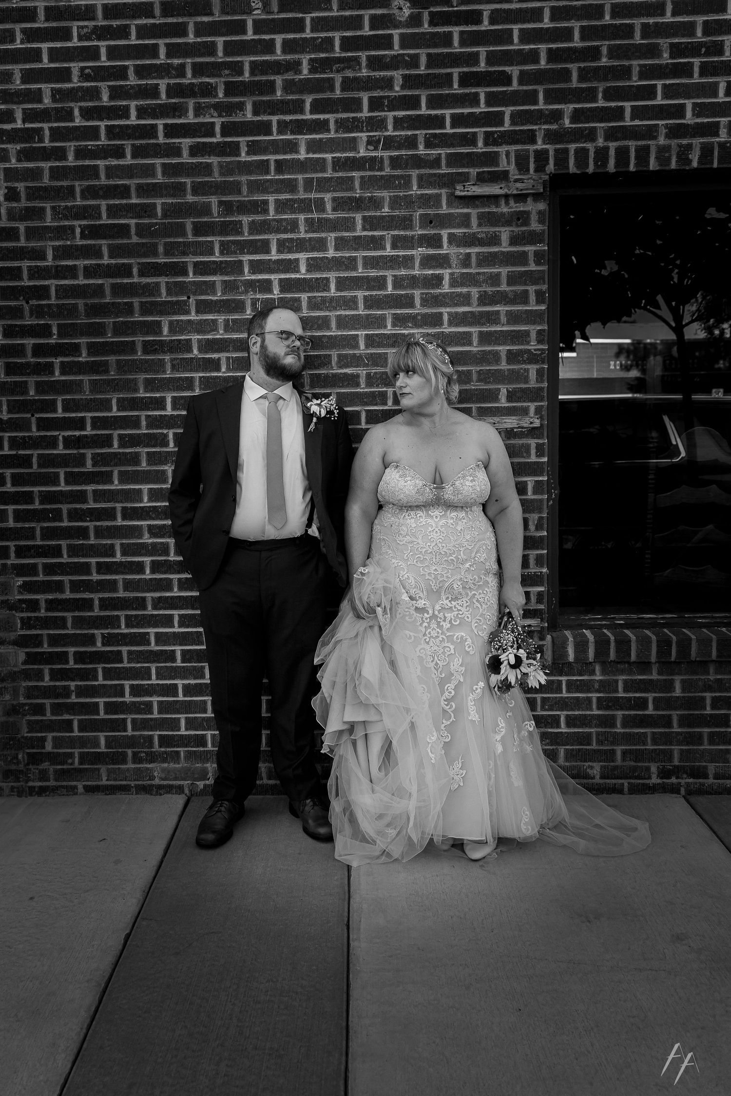 Black and white wedding portrait of a couple standing against a brick wall, with the bride in a lace gown holding flowers.