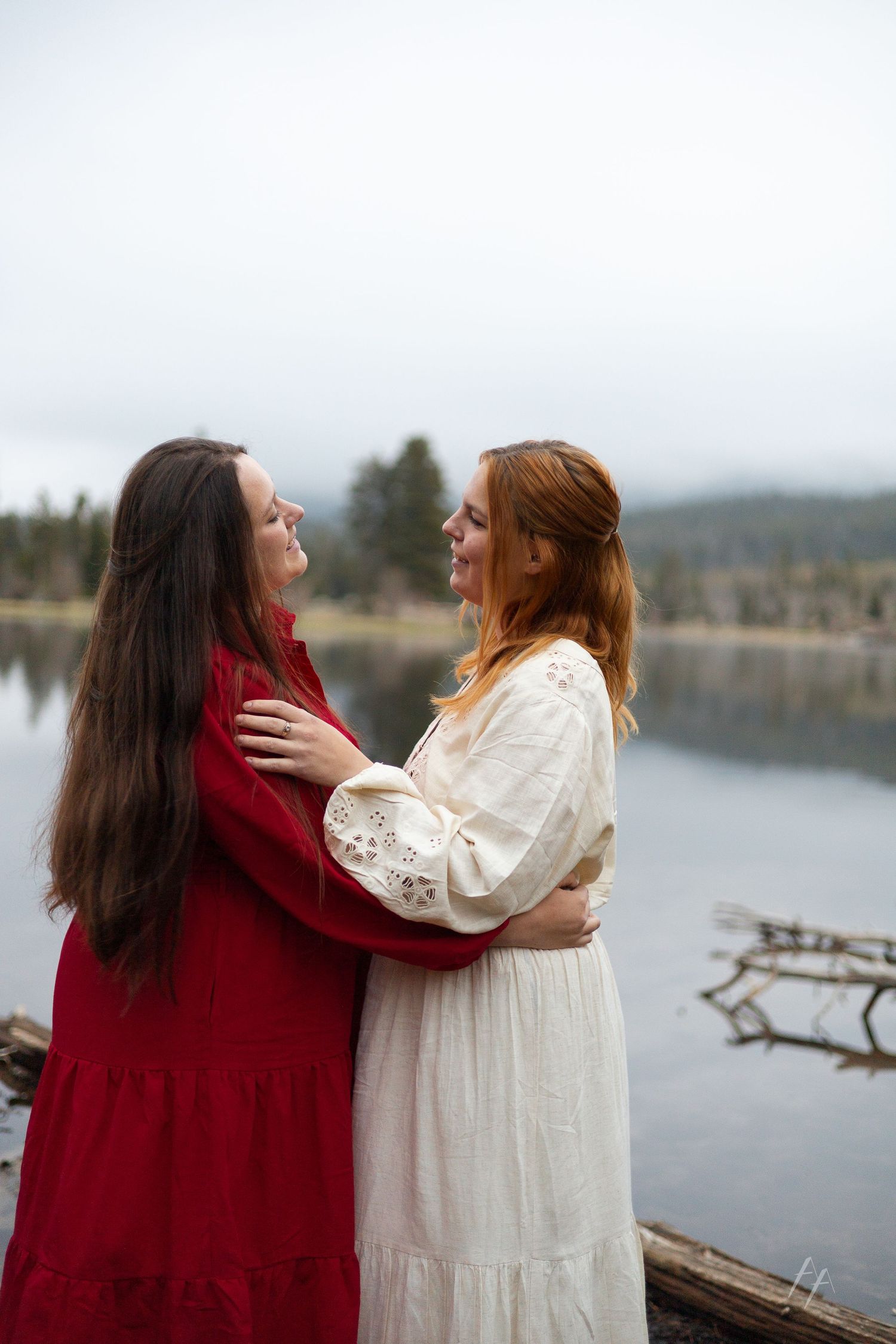 Two women in elegant dresses embrace by a misty lake, one in red and one in white, smiling at each other.