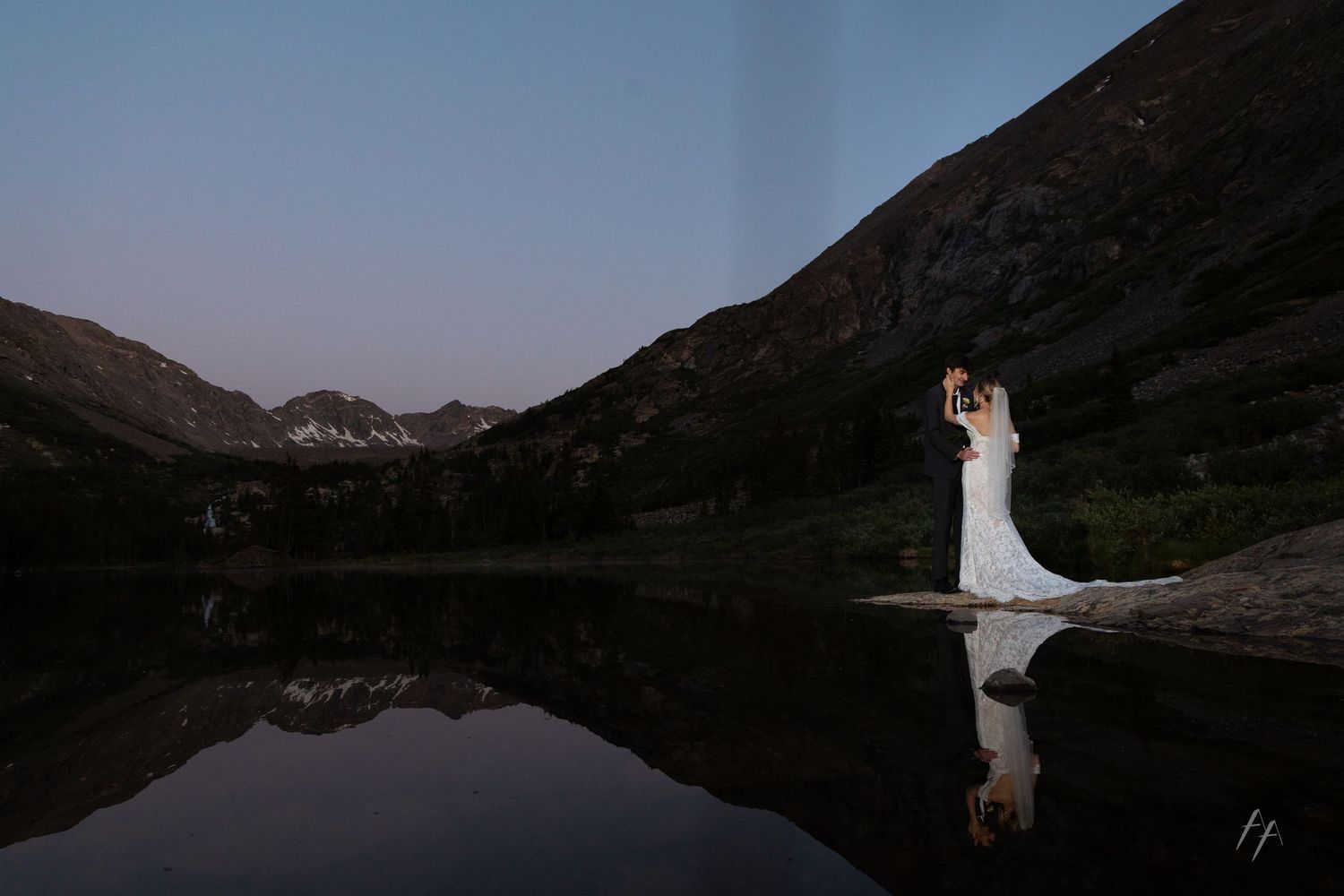 Bride and groom share a kiss by a reflective mountain lake at dusk in a stunning Colorado elopement photo.