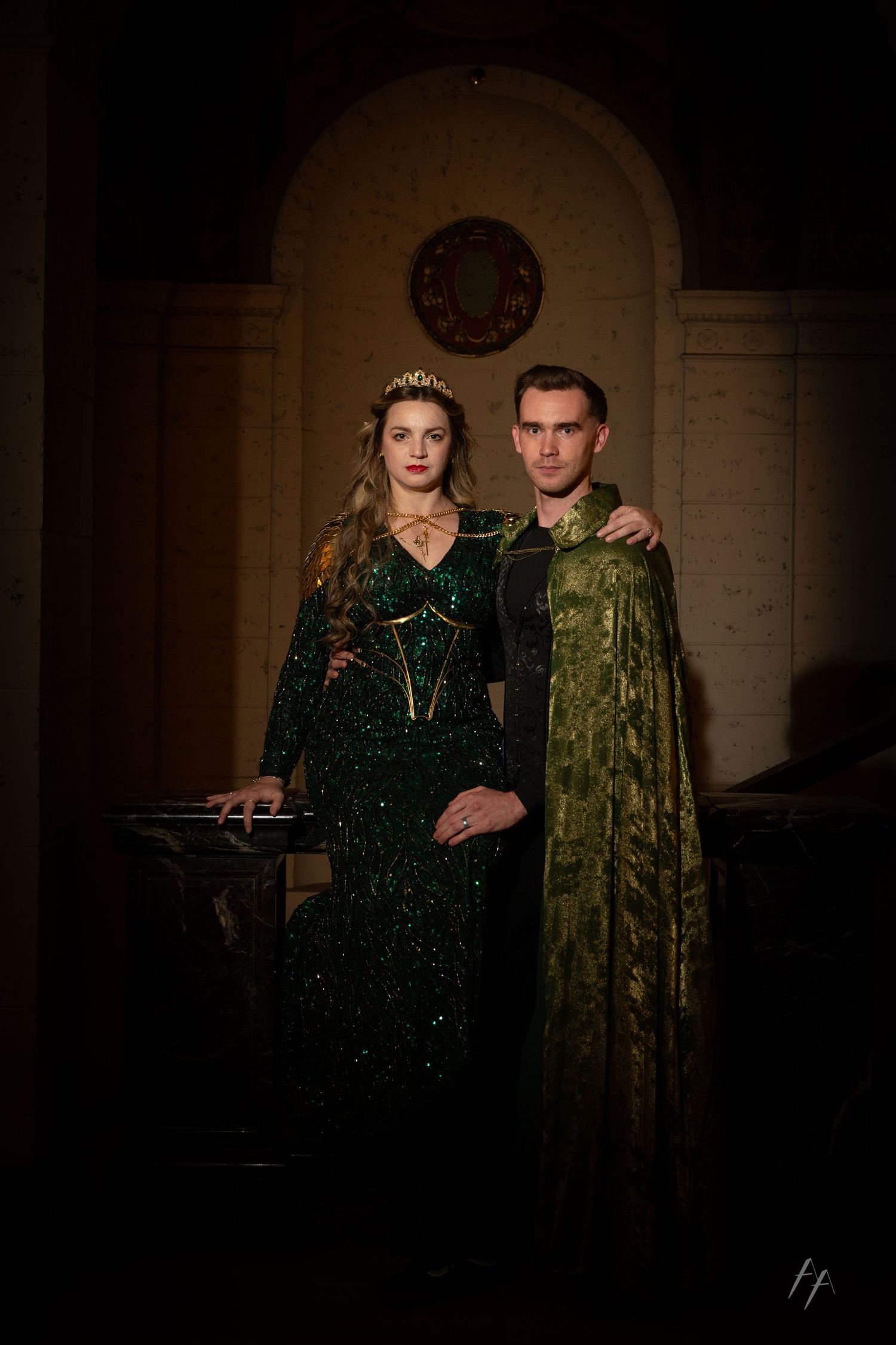A couple poses together in formal attire, with one wearing a dark sequined dress and the other in a green velvet suit.