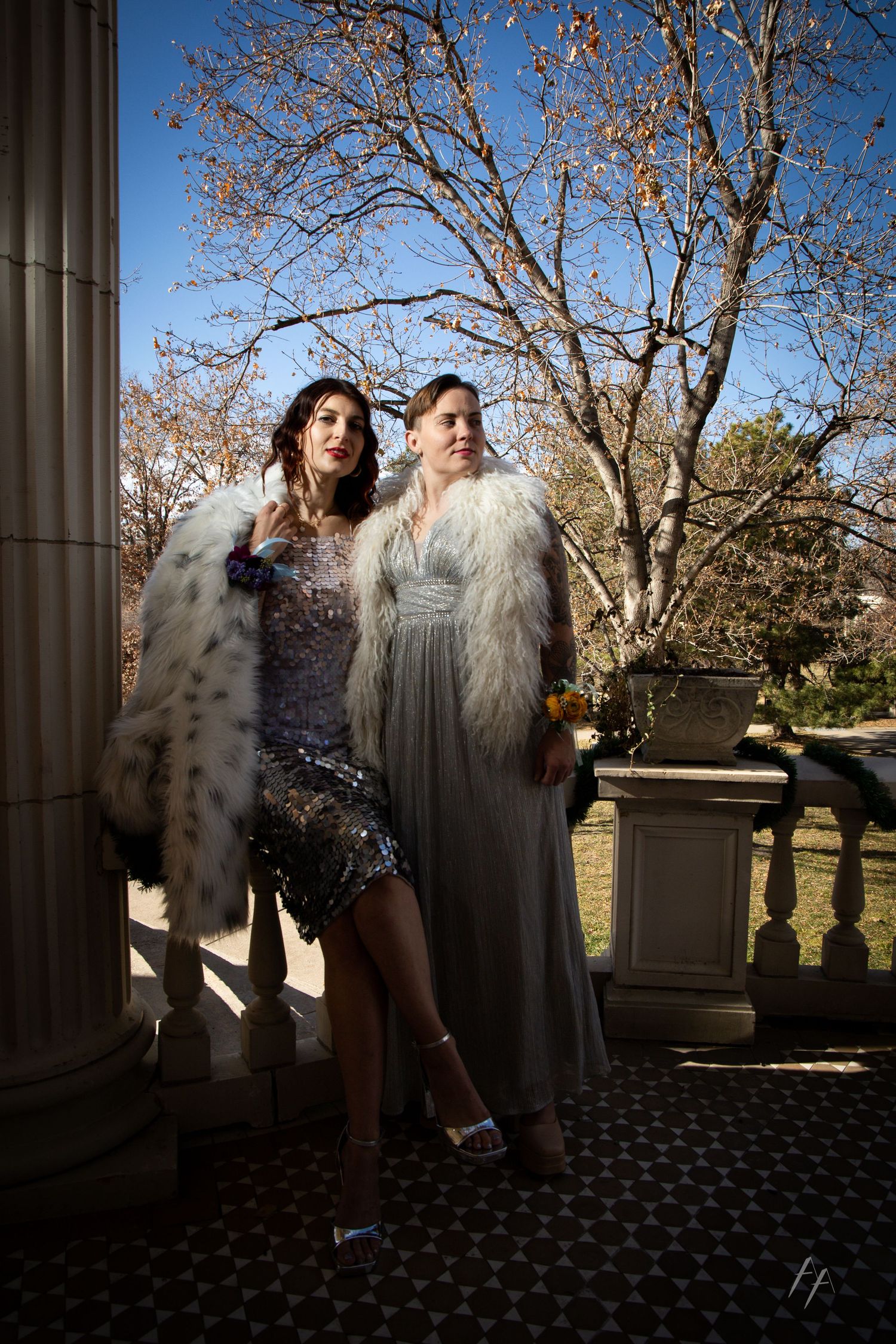 Two people in glamorous evening wear and fur coats pose together against a backdrop of autumn trees.