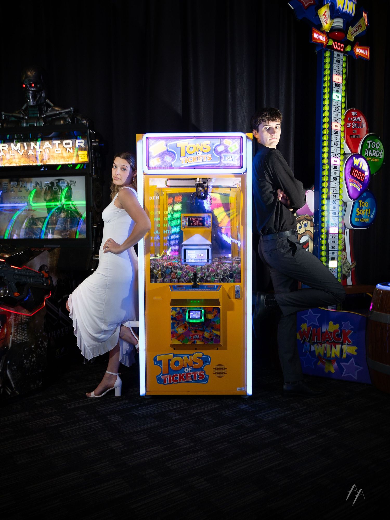 A bride and groom pose back-to-back against a yellow arcade claw machine in a dark game room.