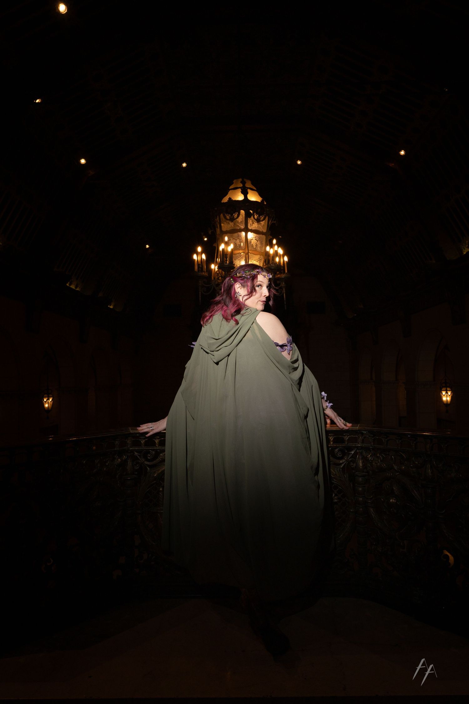 A figure in a flowing sage green cloak stands dramatically backlit in a dark gothic hallway at night.
