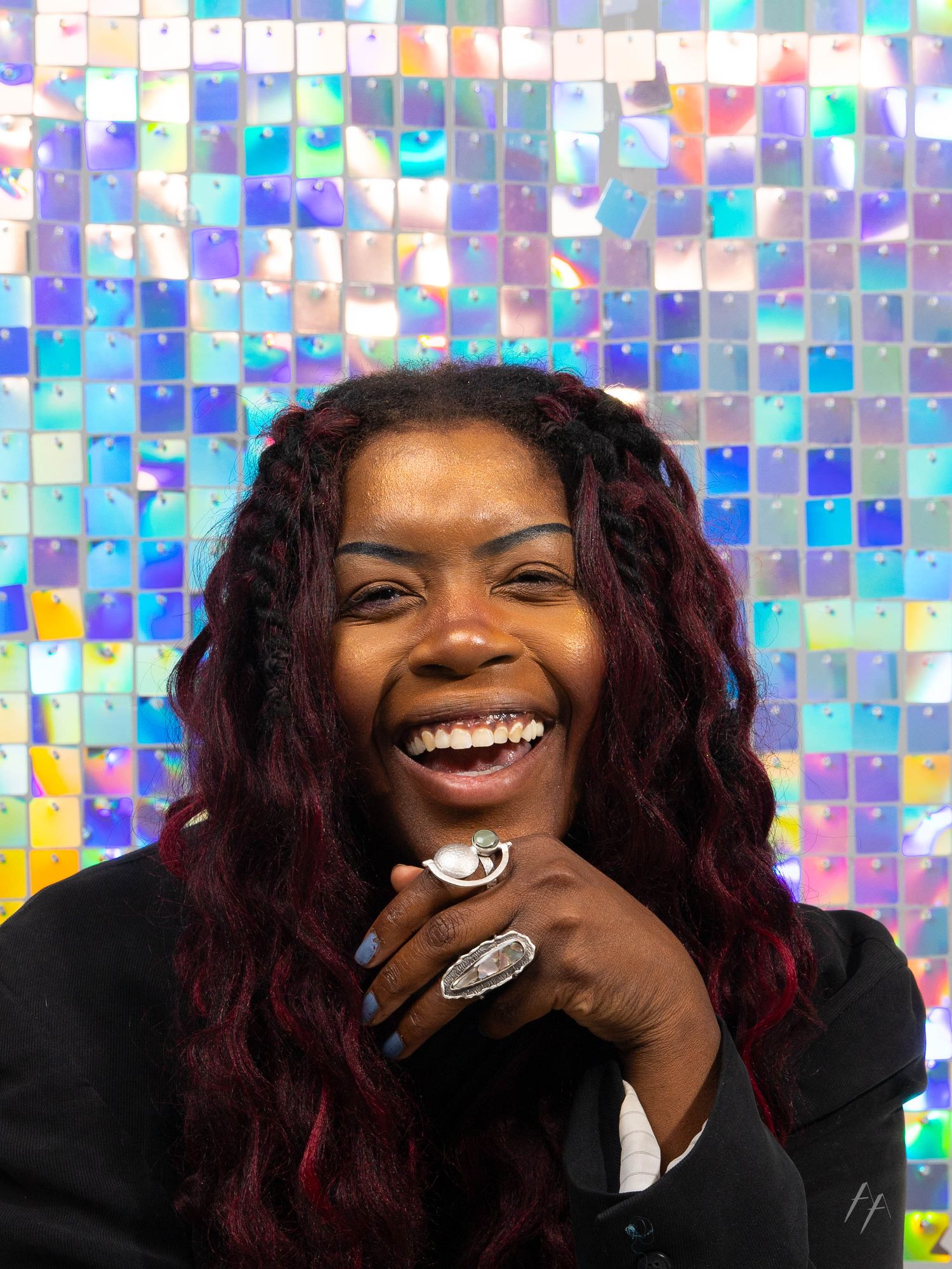 A person with burgundy hair laughs joyfully against a shimmering holographic backdrop while wearing a black outfit.