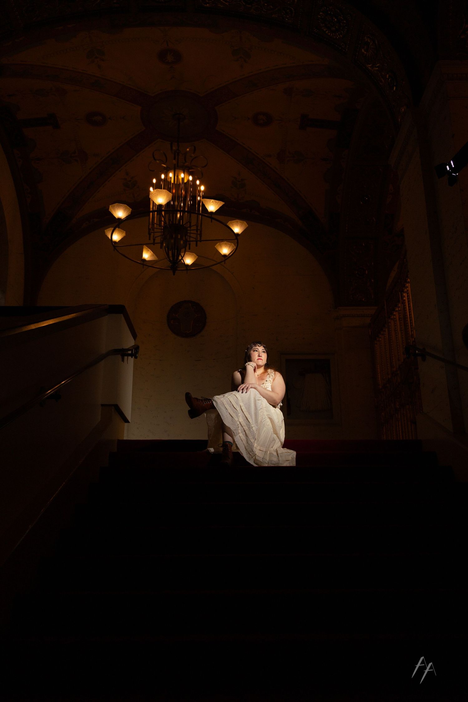 A dramatic low-lit interior scene with a vintage chandelier illuminating a person in a white dress on stairs.