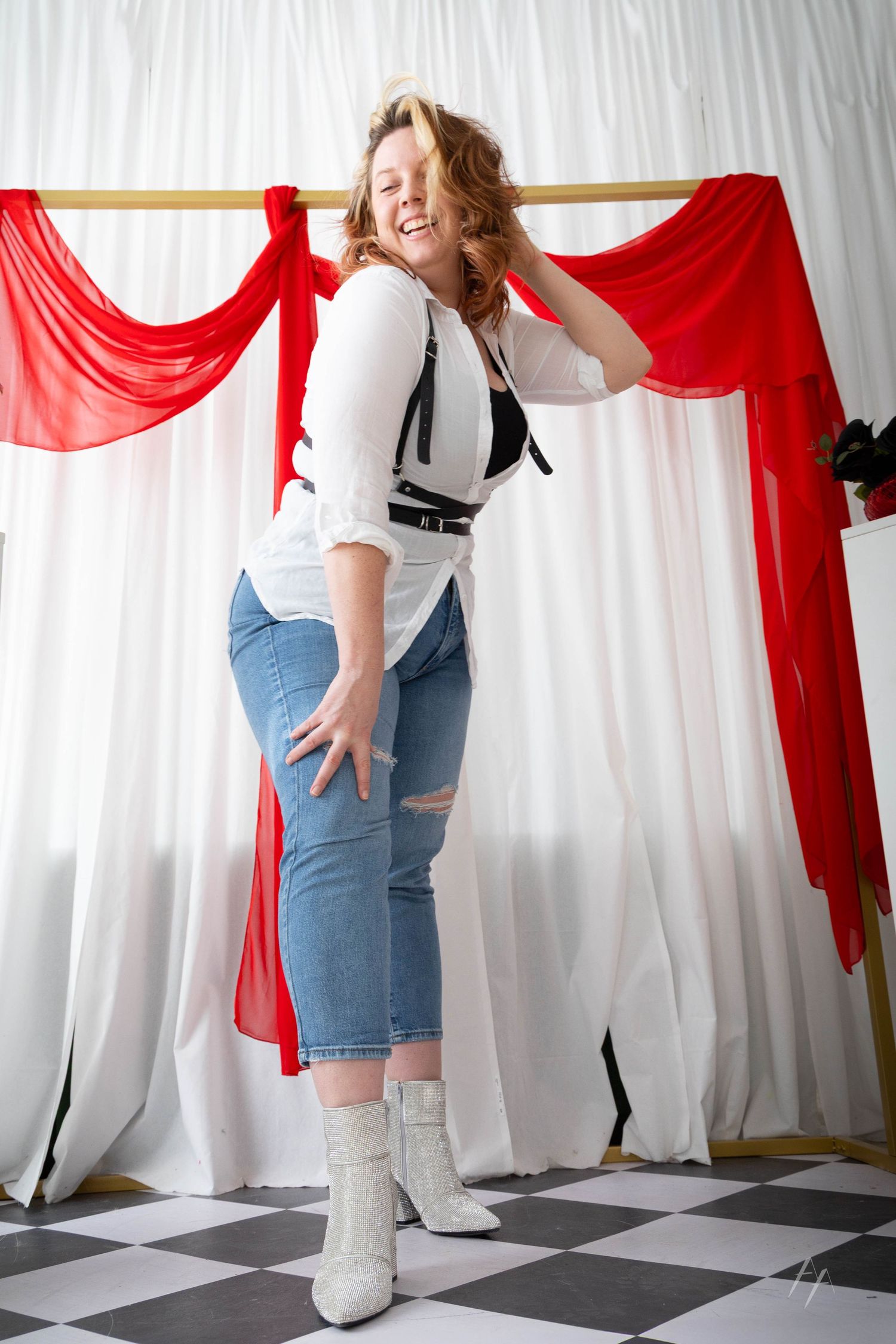 Fashion model in white top and ripped jeans poses against backdrop with red and white draping on checkered floor.