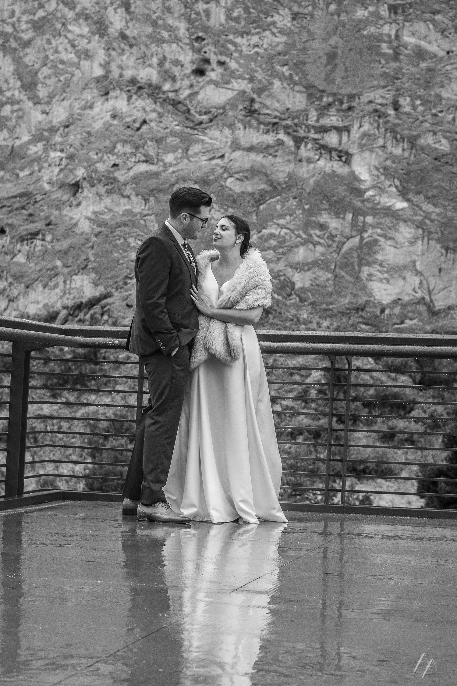 A romantic black and white wedding photo of a couple sharing an intimate moment on a rainy deck with stone wall backdrop.
