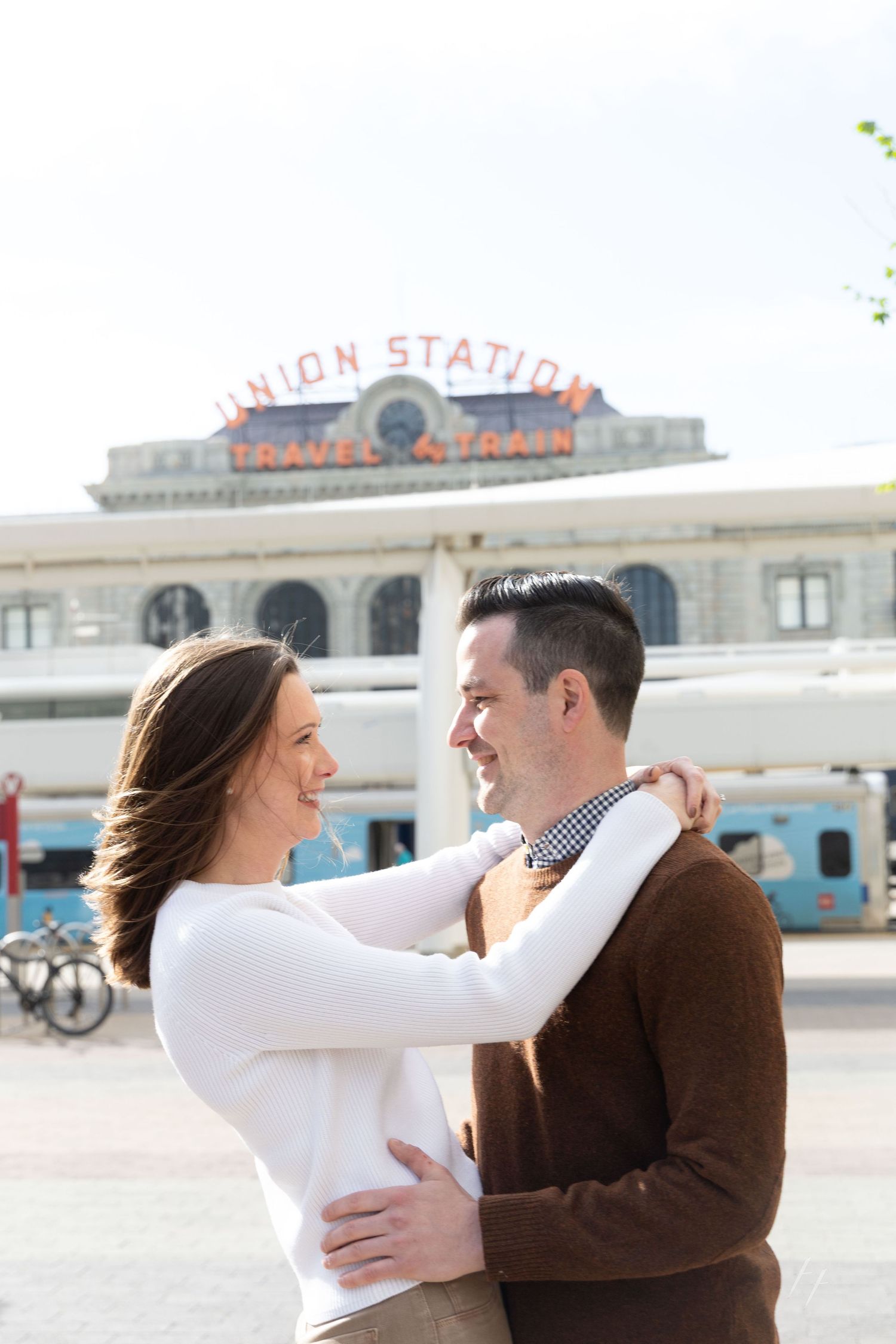 A couple embraces and smiles at each other in front of Denver Union Station's iconic travel and train sign.