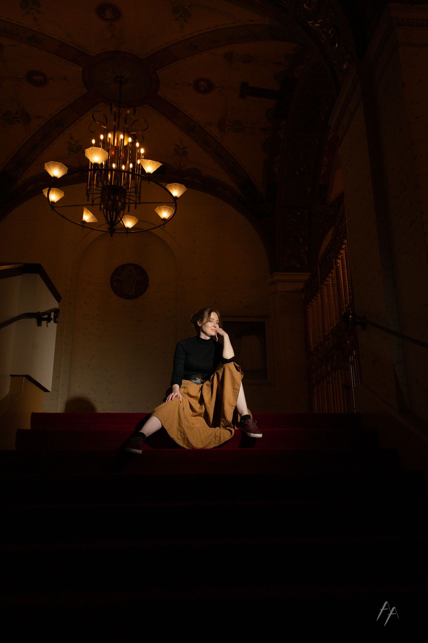 A dimly lit concert hall interior with elegant chandeliers and arched ceilings creates a dramatic atmospheric ambiance.