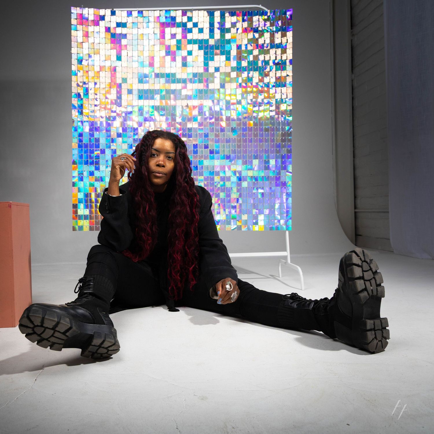 Figure in black outfit with chunky boots sits on white floor against pixelated digital art backdrop.