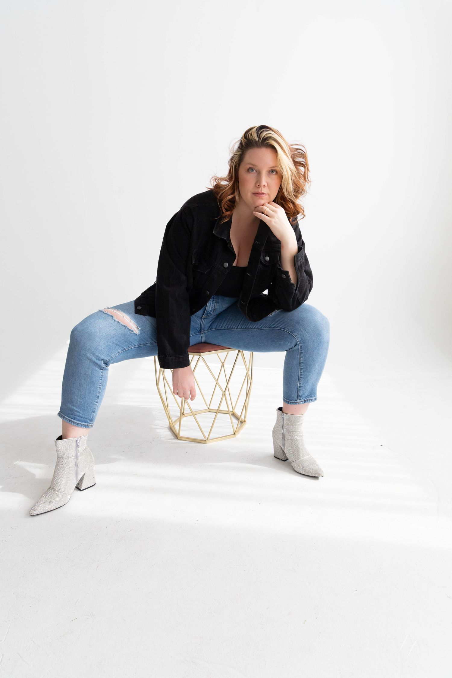 Person in black blazer and light jeans poses artistically with geometric gold stool against white backdrop.